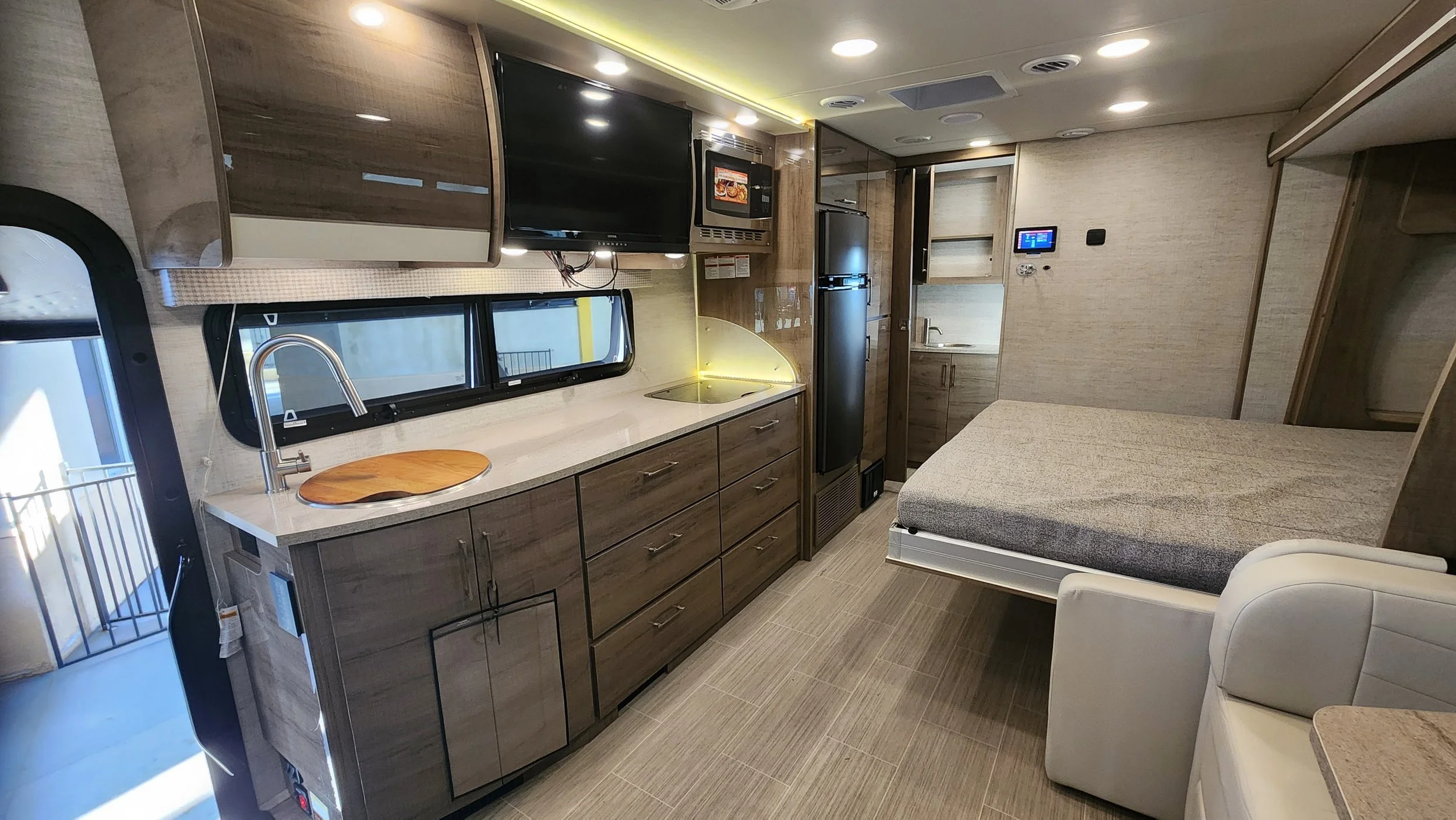 Interior of a compact RV with kitchen appliances, a window, a bed, a small couch, and built-in cabinets.