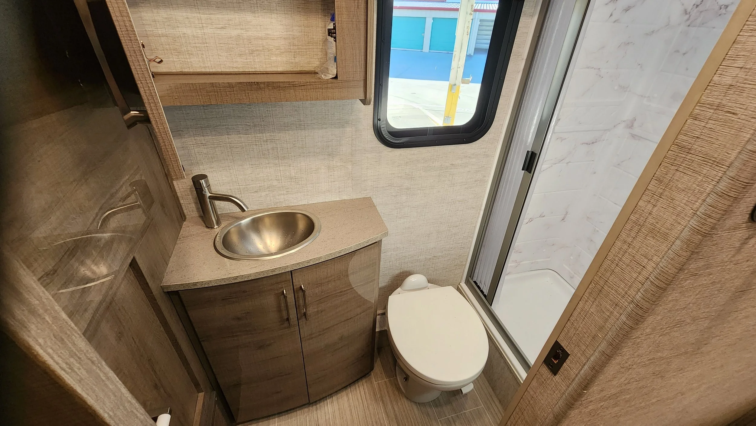 Small bathroom with a corner vanity and stainless steel sink, toilet, and enclosed shower with sliding door. A window is above the sink, showing an outdoor area with blue siding. The walls and floor have a wood and marble appearance.