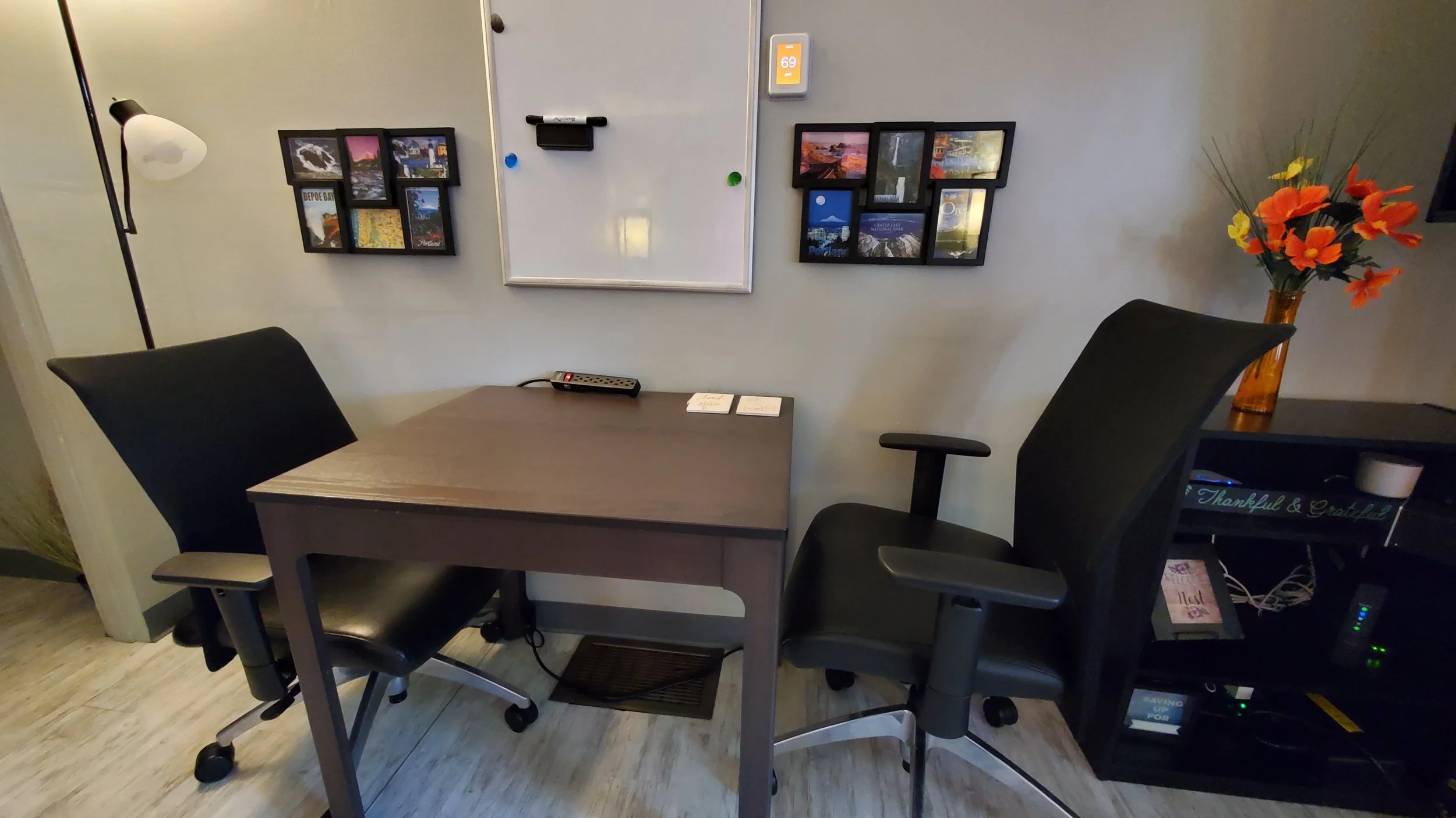 Home office or study space with a wooden desk, two black office chairs, a whiteboard on the wall, framed photographs, a vase with orange and yellow flowers, and a black shelf with books and electronics.