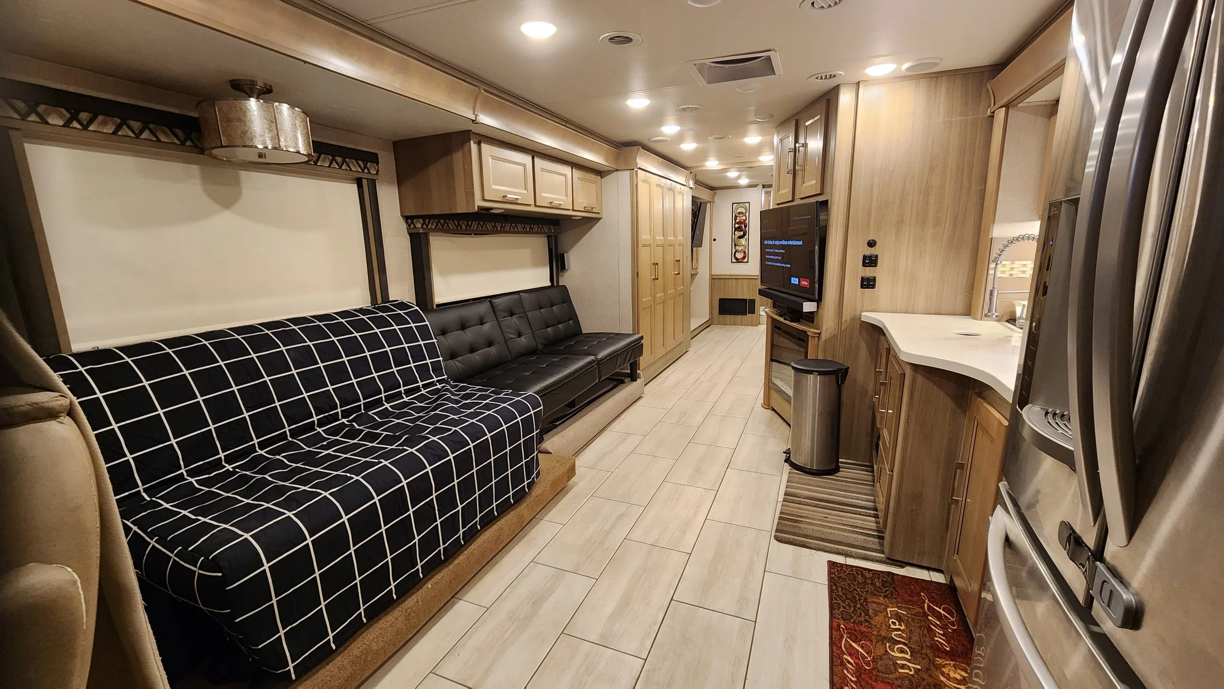 Interior of a RV living area with beige walls, wooden cabinetry, two black sofas, a flat-screen TV, a small kitchenette with a white countertop, and a stainless steel refrigerator.