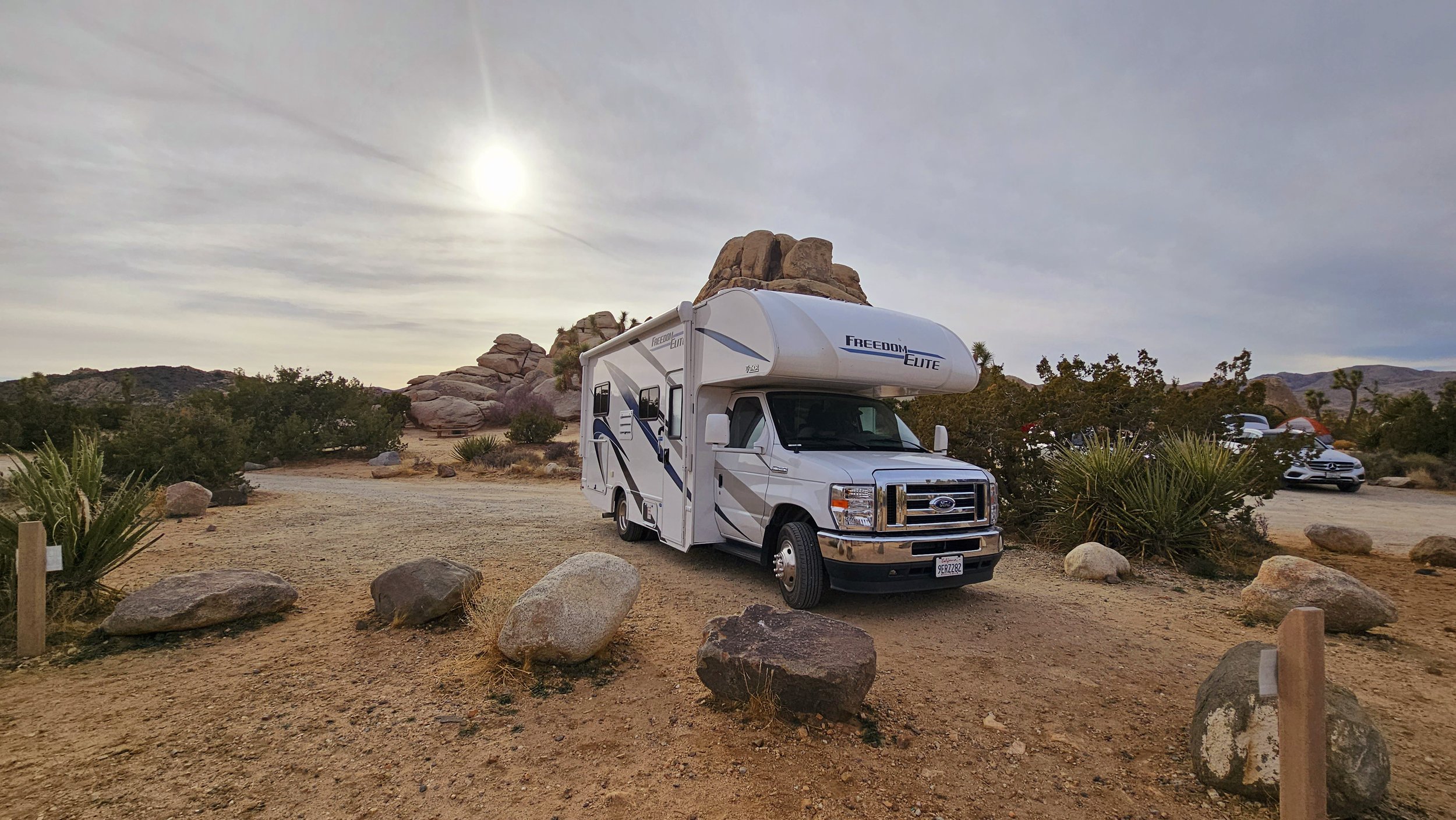 A white recreational vehicle (RV) parked in a desert landscape with rocks, bushes, and distant mountains under a cloudy sky.