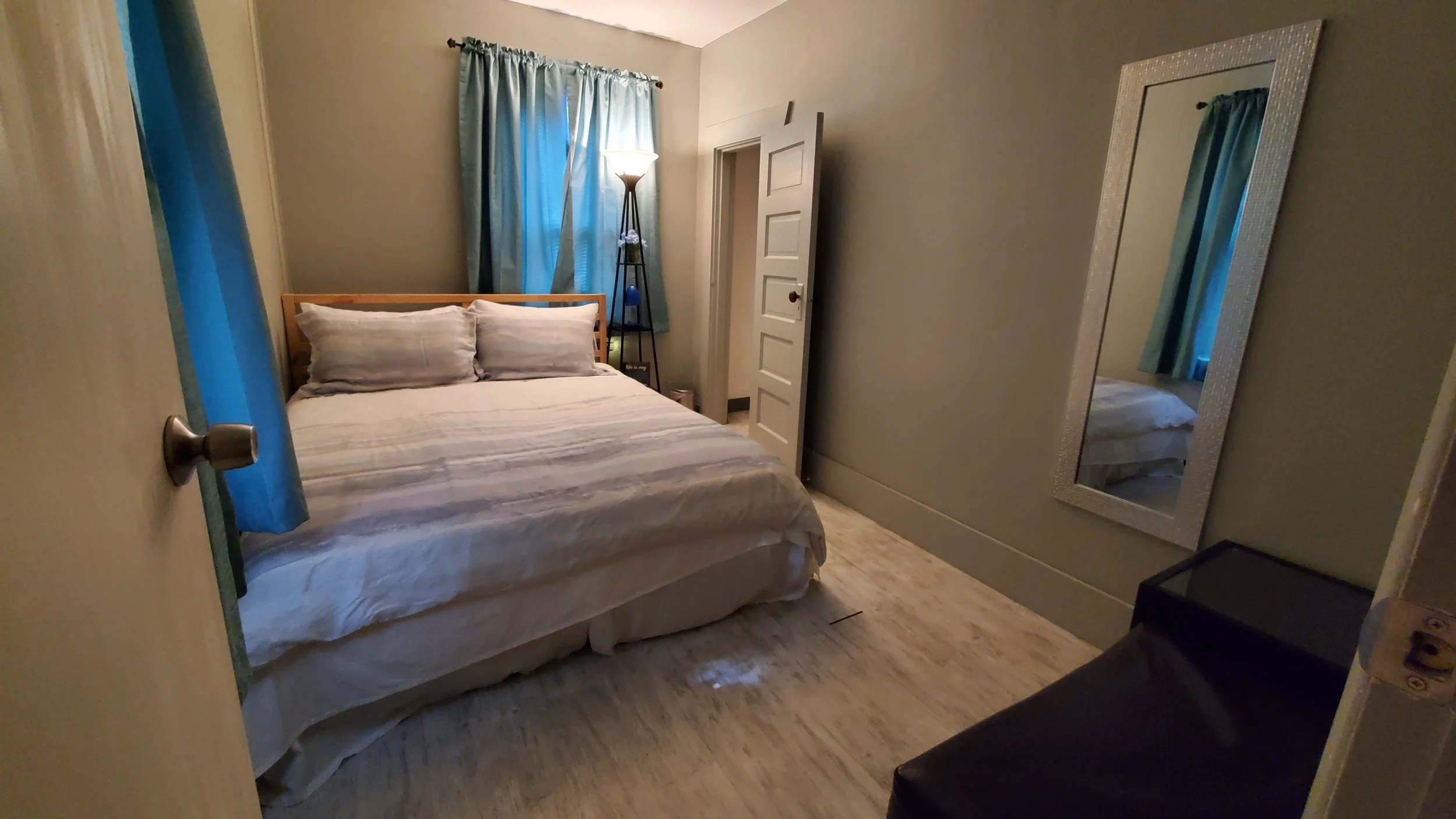 A cozy bedroom with a bed, a mirror on the wall, a window with blue curtains, a lamp, and a small black table.