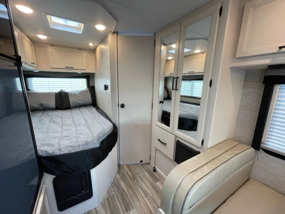 Interior of a compact mobile home or RV with a bed, mirror-front cabinets, a window, and a small sitting area.