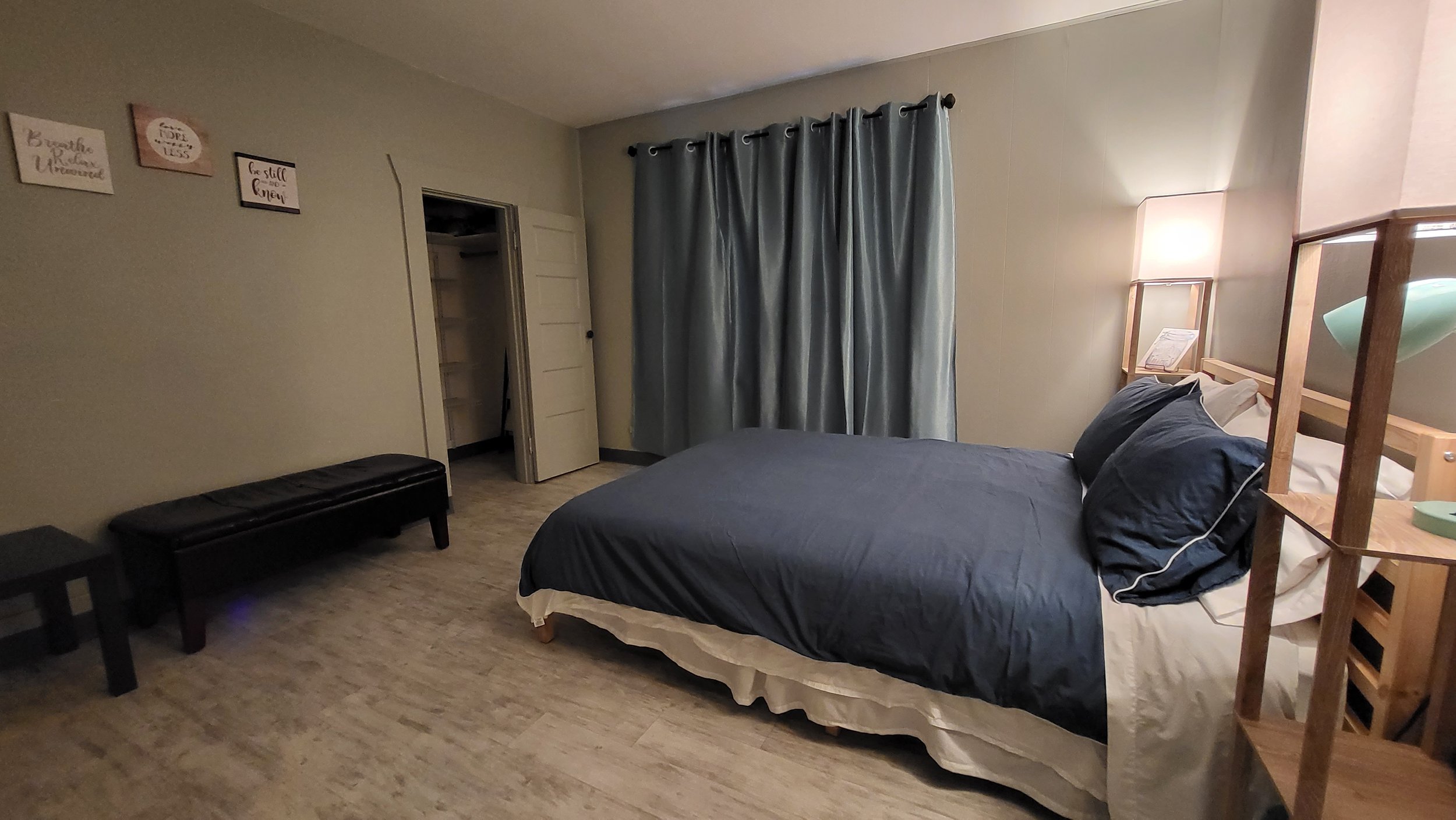 A bedroom with a bed covered in dark blue bedding, a wooden headboard with built-in shelves, a nightstand with a lamp, gray curtains on a window, a closet with open doors, and decorative wall hangings.