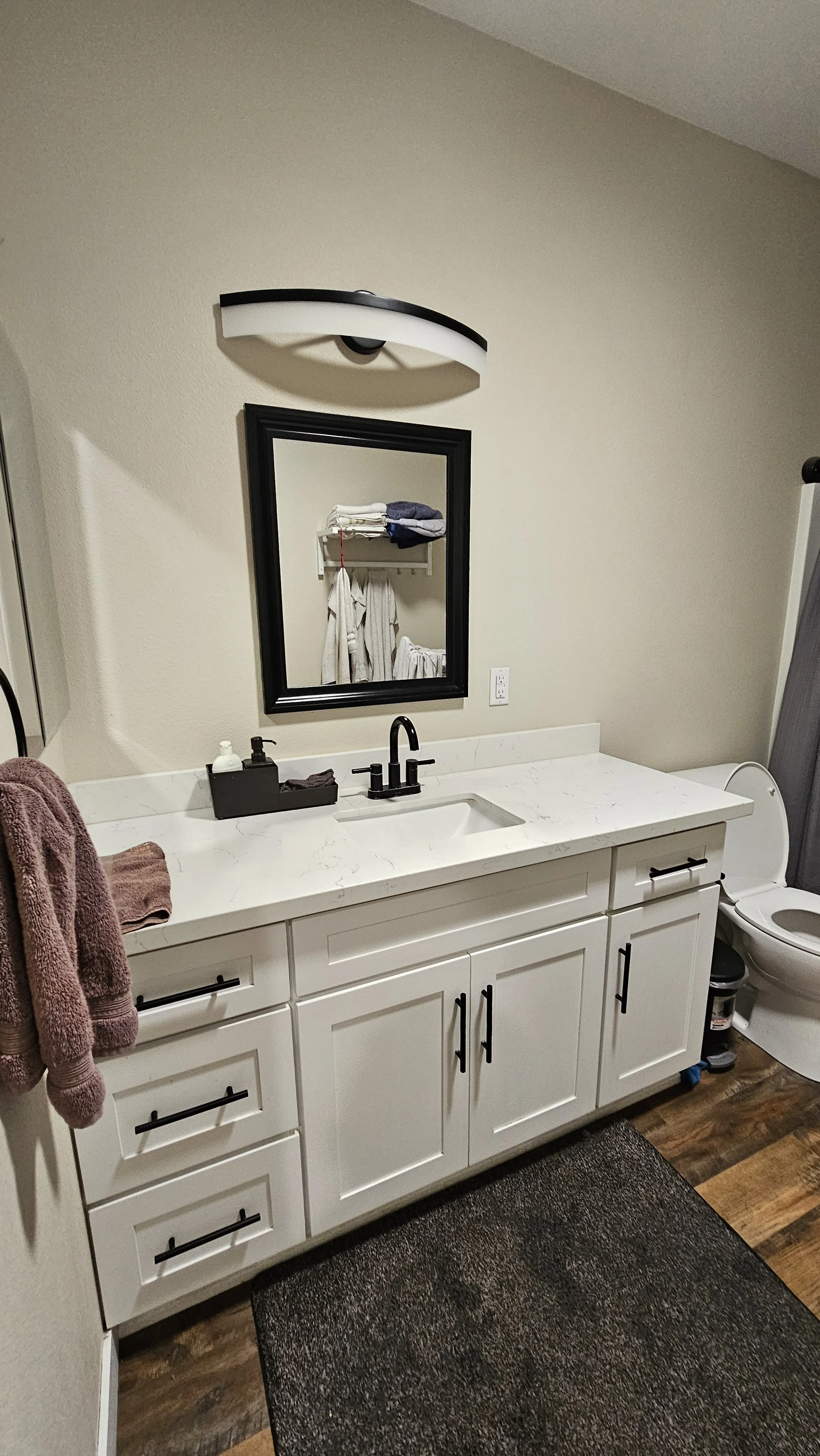 Bathroom vanity with white cabinets, black handles, a black-framed mirror, a white marble countertop, a black faucet, a soap dispenser, and a towel hanging on the side. A toilet with an open lid is visible to the right, and a dark bathroom rug is on 