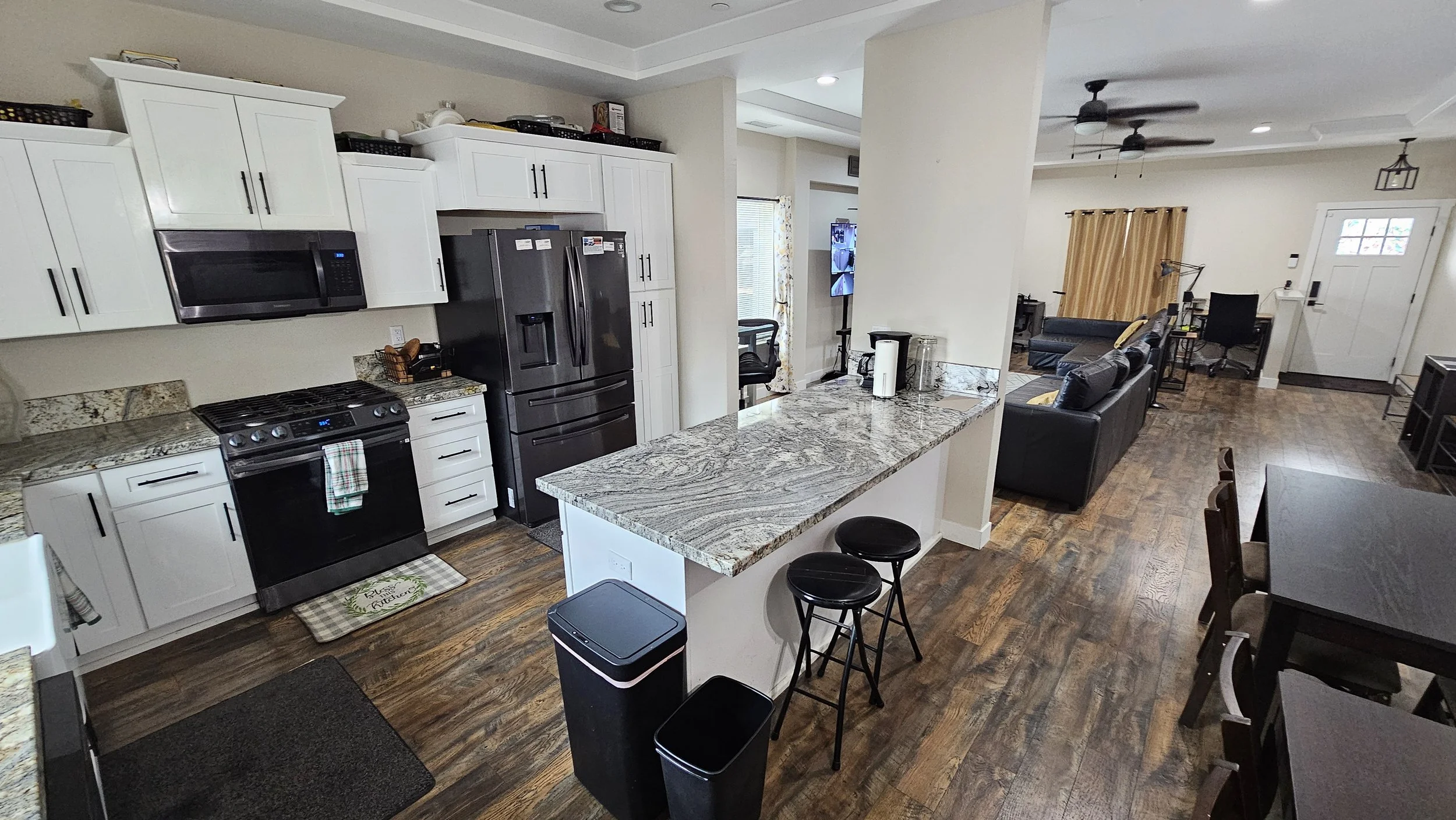 Open-concept kitchen and living room with white cabinets, granite countertops, black appliances, a dining table, and a sitting area with a TV.