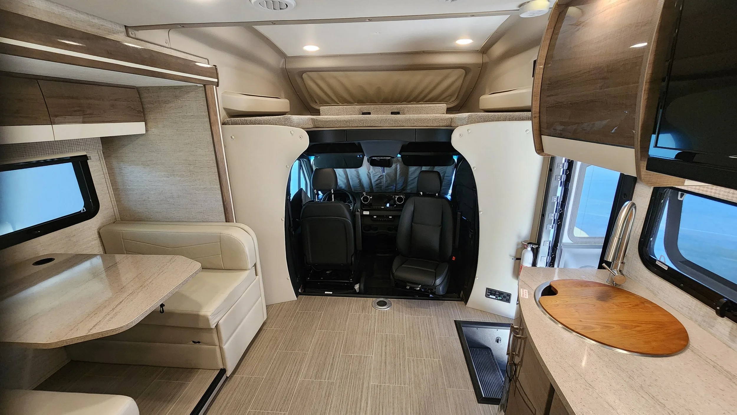 Interior of a motorhome with a small dining area, kitchen with a sink, a cab with two black leather seats, and a sleeping loft above.