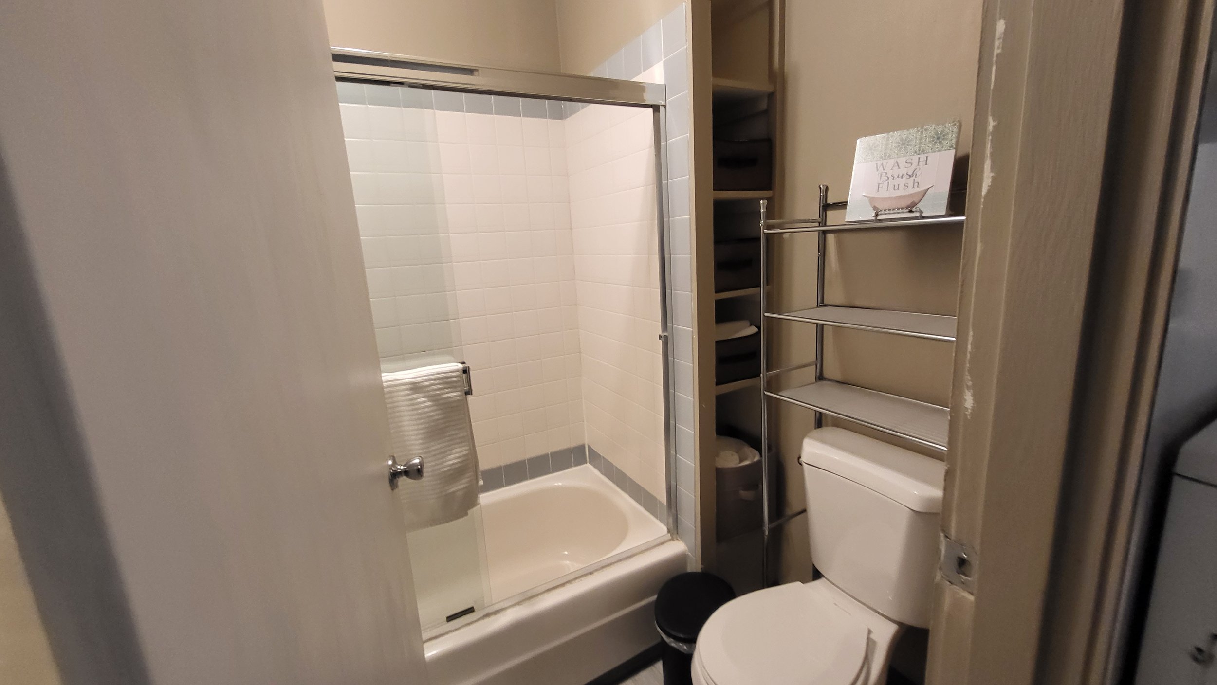 Small bathroom with a bathtub and shower, toilet, shelving unit, and trash bin.