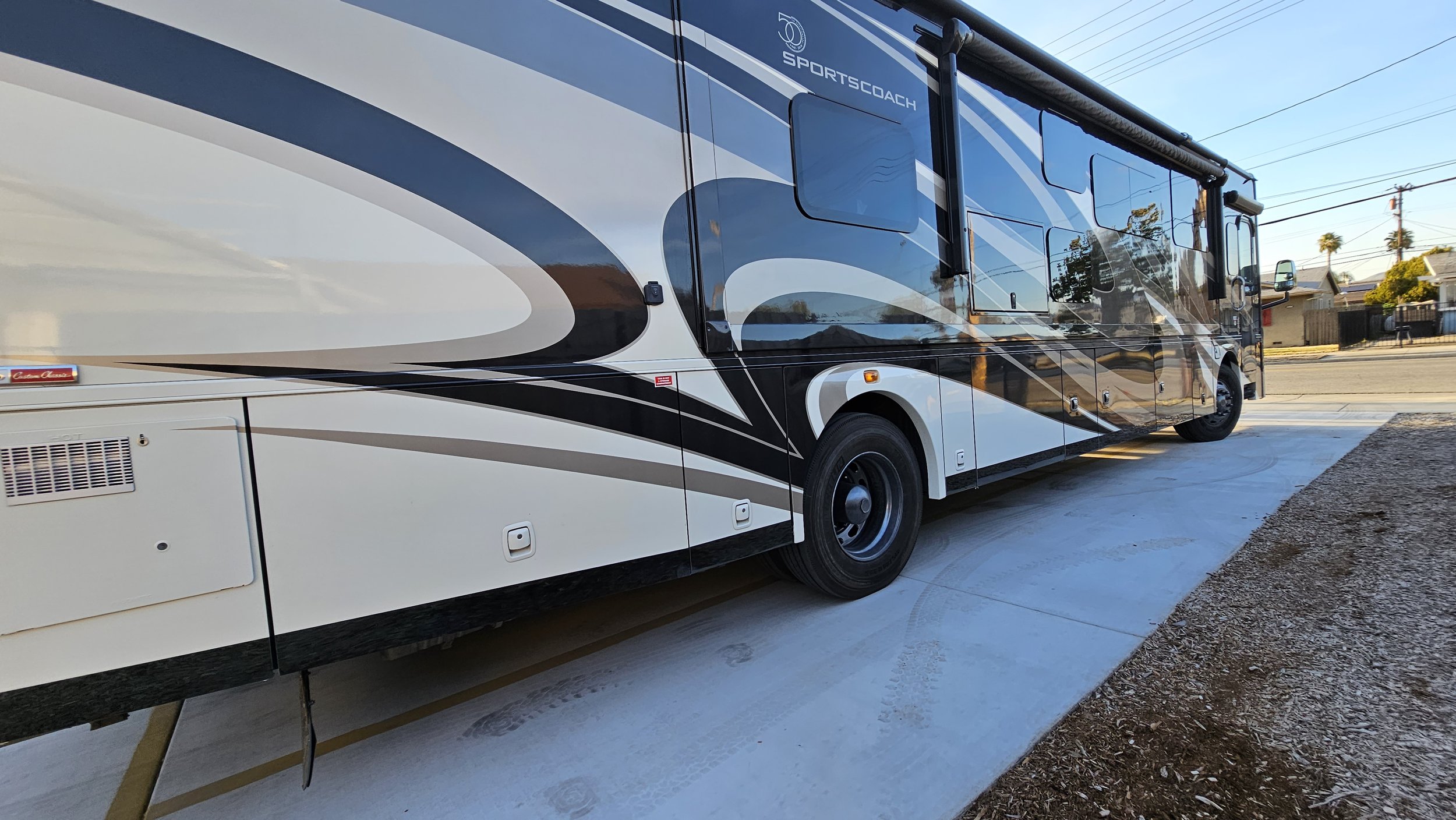 A large white, black, and gray sports coach espresso motorhome parked on a concrete driveway with a residential neighborhood in the background.