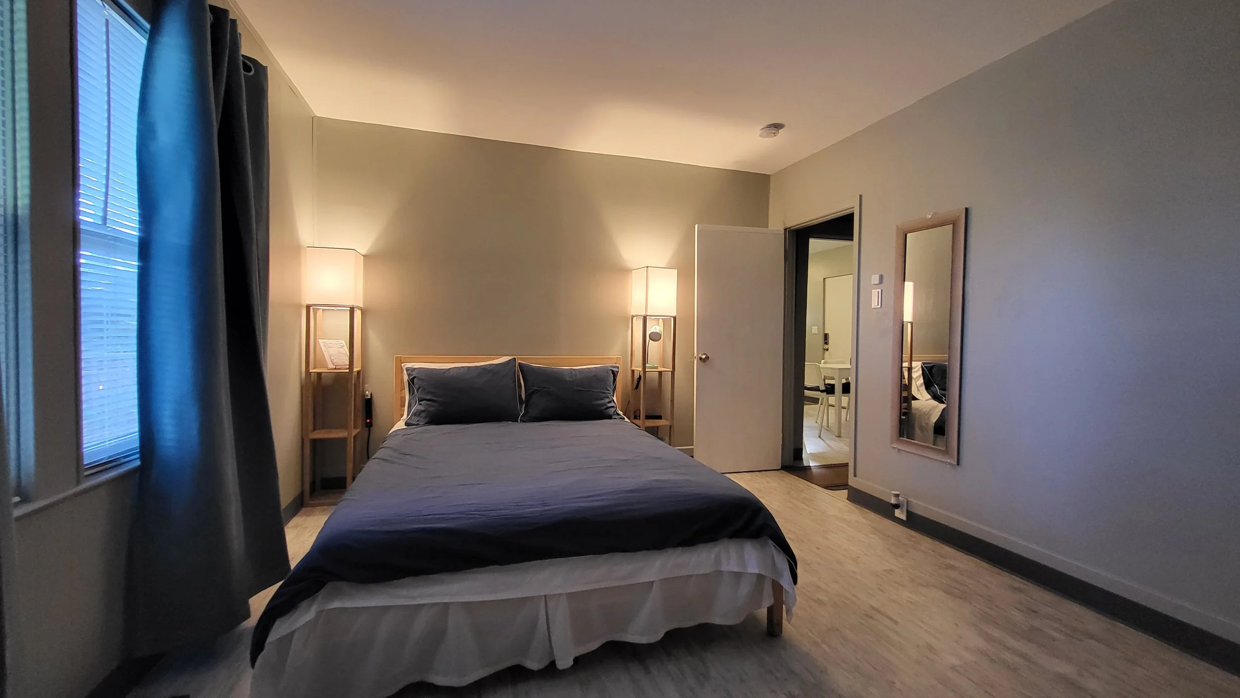 A neatly made bed with dark bedding and pillows in a cozy, modern bedroom with wooden nightstands and lamps. There is a mirror on the wall and an open door leading to another room.