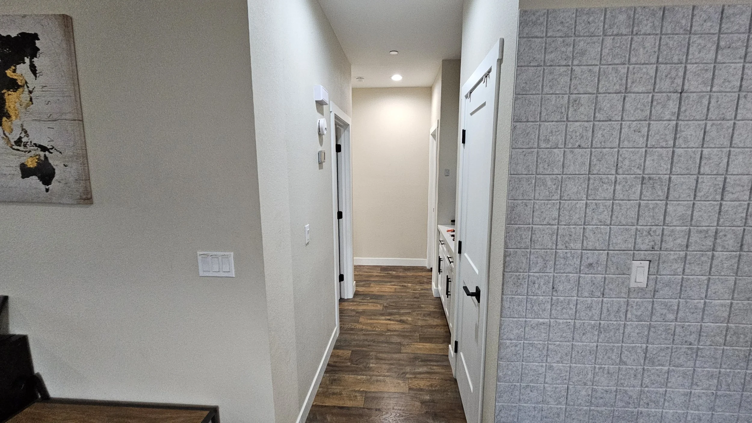 Interior view of a hallway with wooden floor, beige walls, multiple doors, and a textured gray wall on the right side.