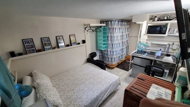 Small studio apartment with a bed, a small kitchen area, shelves, and a privacy curtain.
