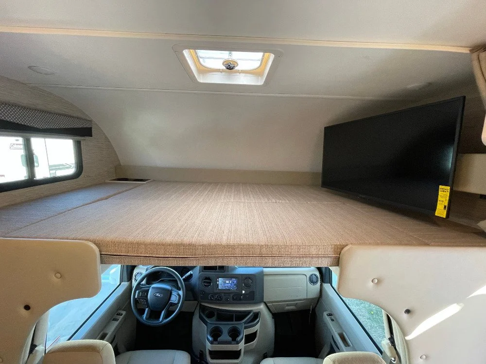 Interior of an RV, showing a bed space above the driver's cockpit with a small TV on the right side and a window on the left side.