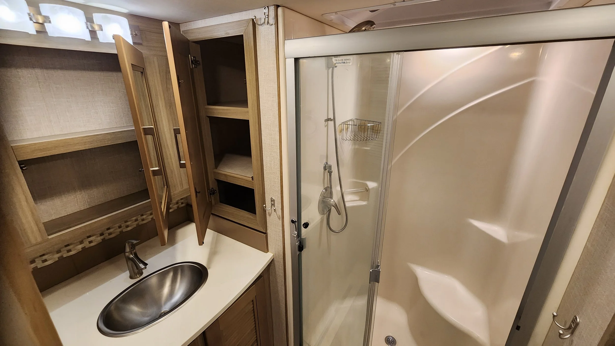 Small bathroom with a wooden cabinet, a shower with a glass door and built-in shelves, a white counter with a stainless steel sink, and beige walls.