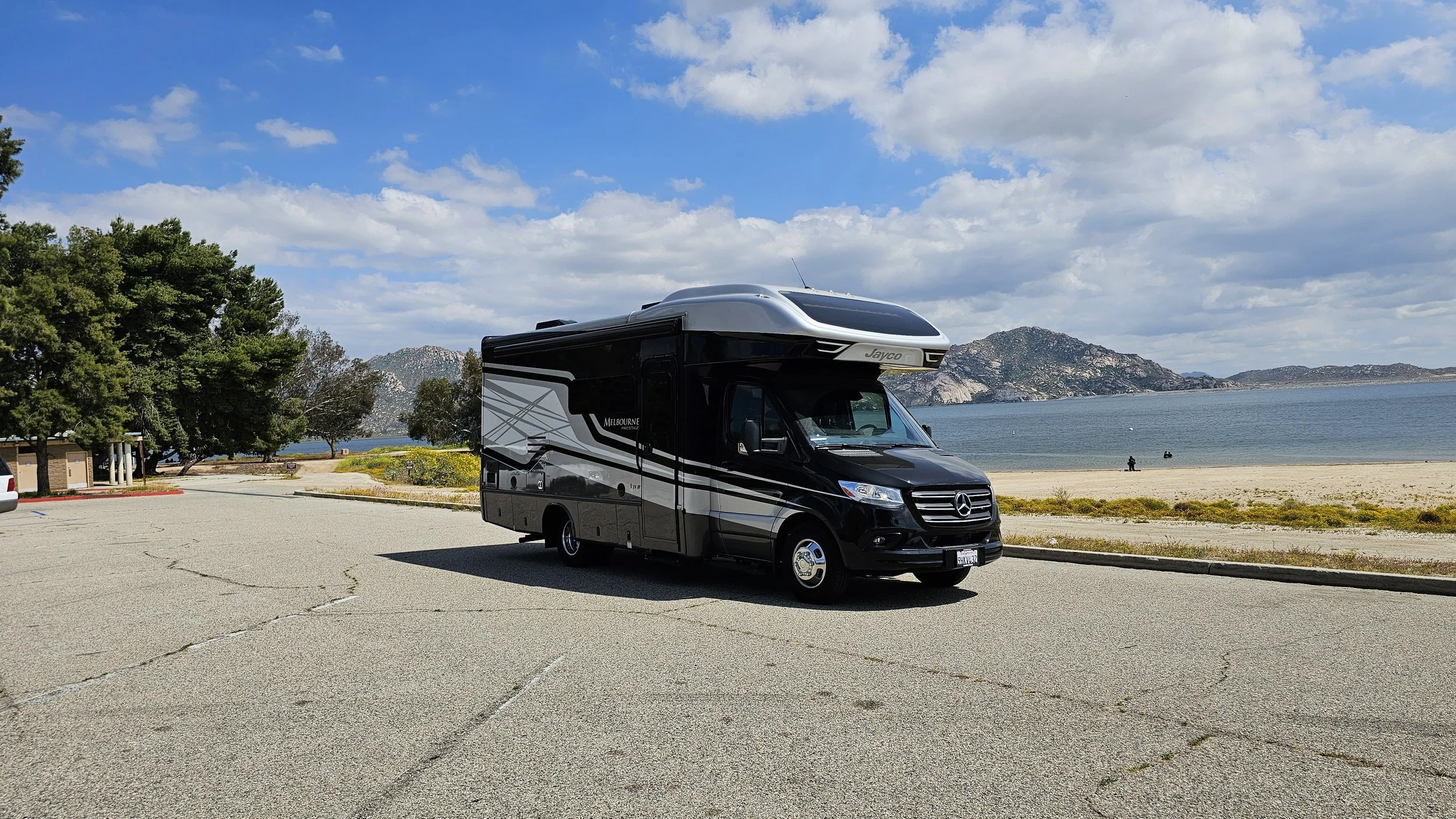 Black and gray RV parked near a beach with mountains and water in the background, under a partly cloudy sky.