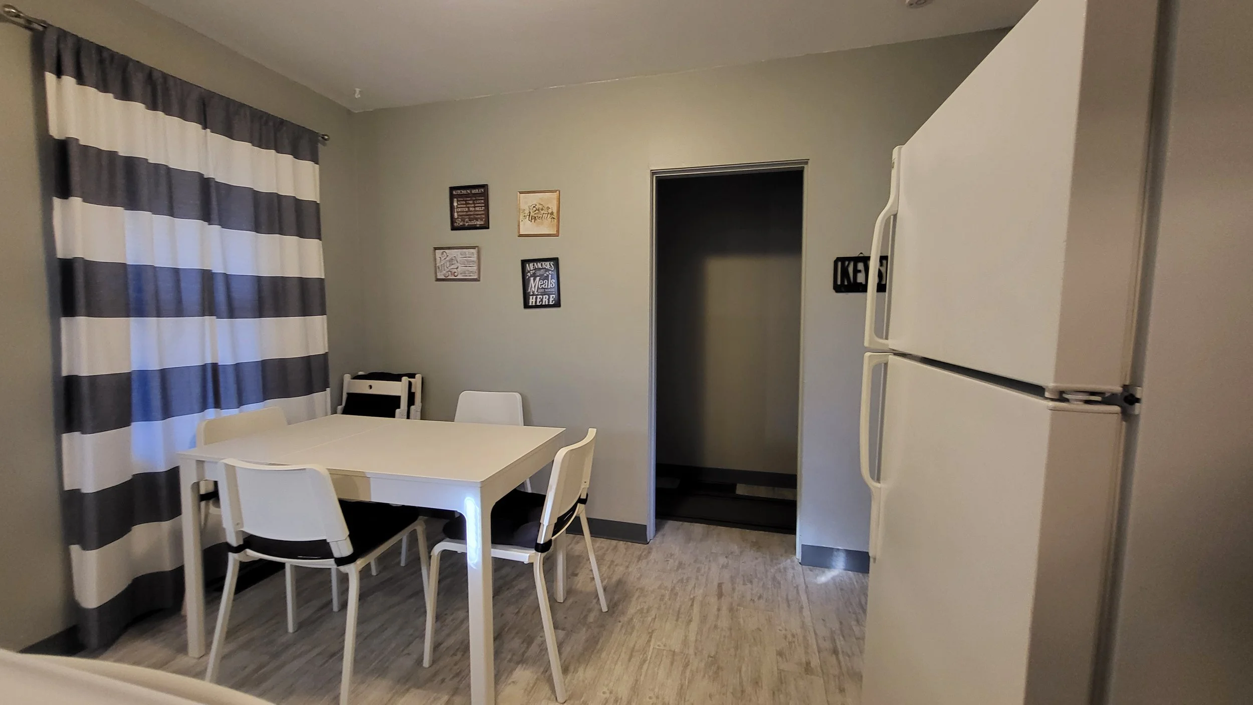 Dining area with a white table, four chairs, a window with black and white striped curtains, wall decorations, and a white refrigerator.