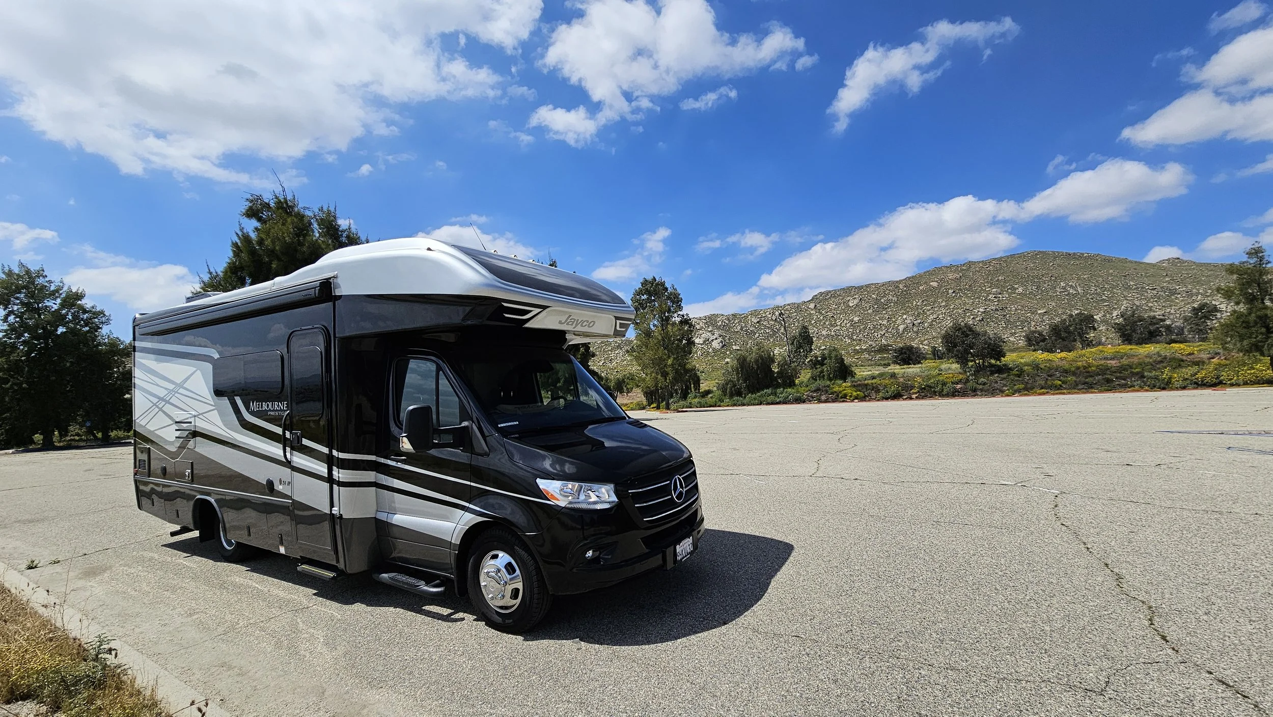 Black Mercedes-Benz motorhome parked in an empty lot with mountains and blue sky with scattered clouds in the background.
