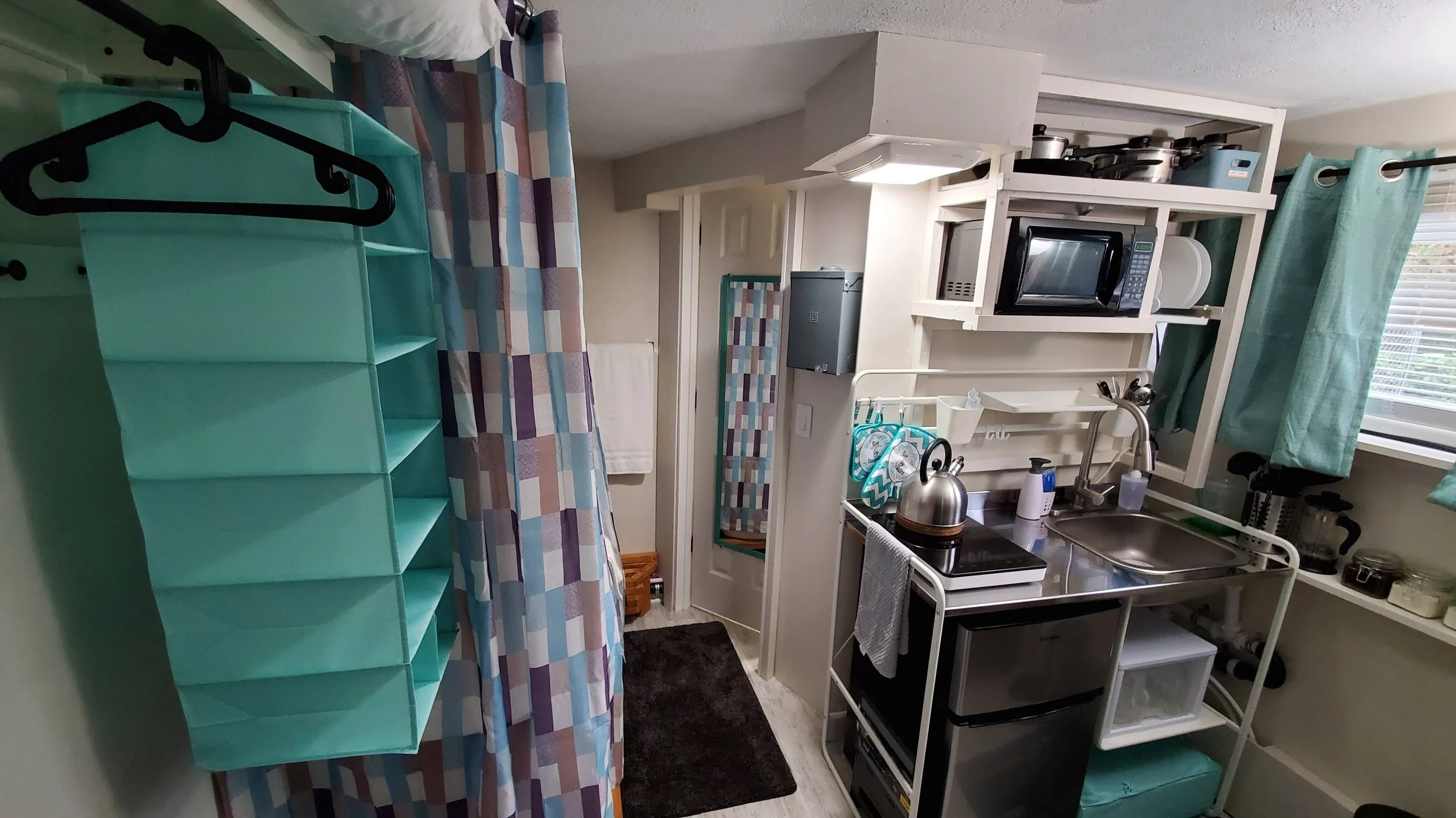 Compact kitchen area with microwave, small fridge, sink, and storage shelves. Clear turquoise curtains, patterned shower curtain, and black rug on wooden floor.