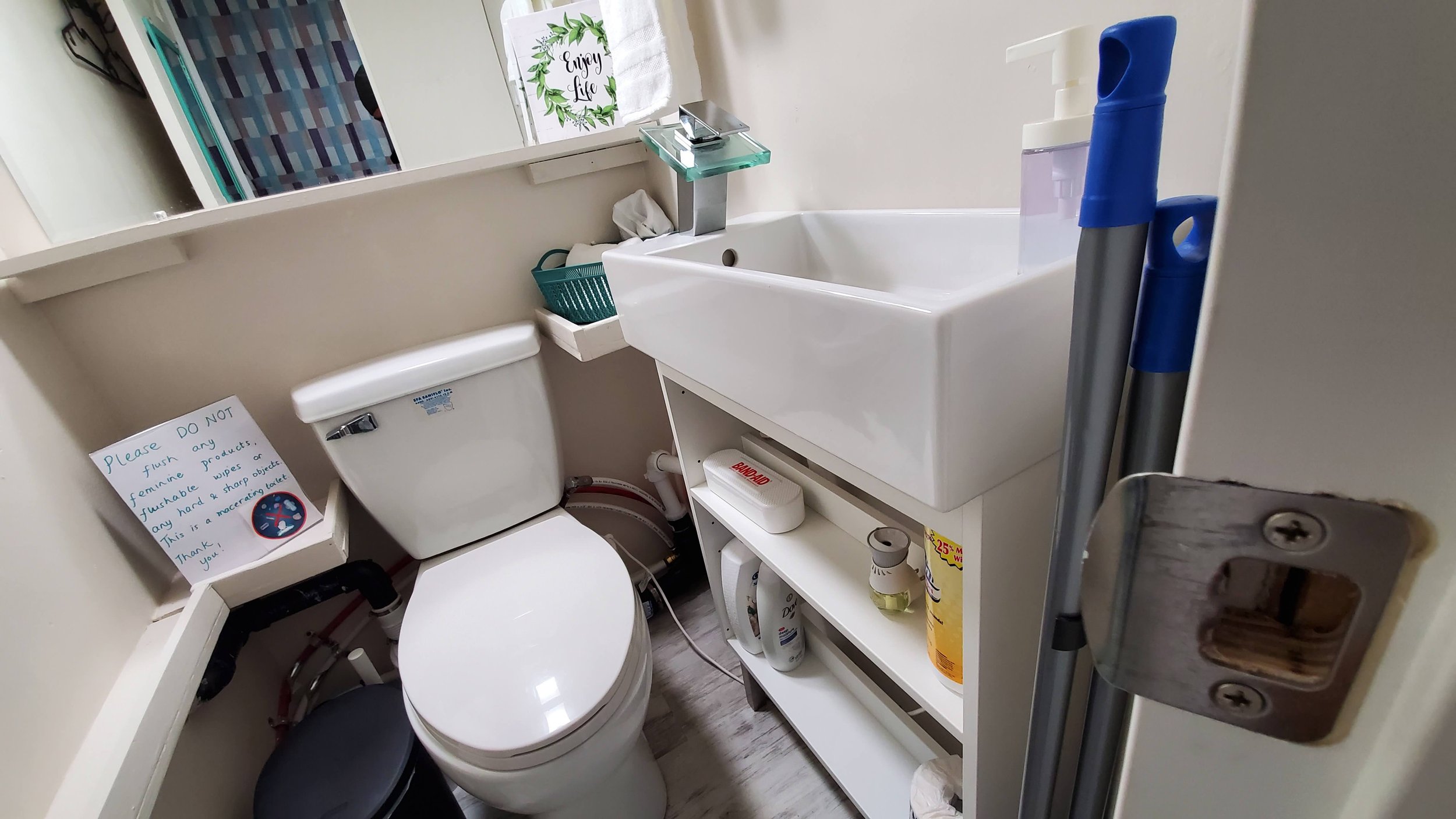 Small bathroom with a toilet and a white sink, cleaning supplies, a mirror, and a sign on the toilet with a note about not flushing feminine products.