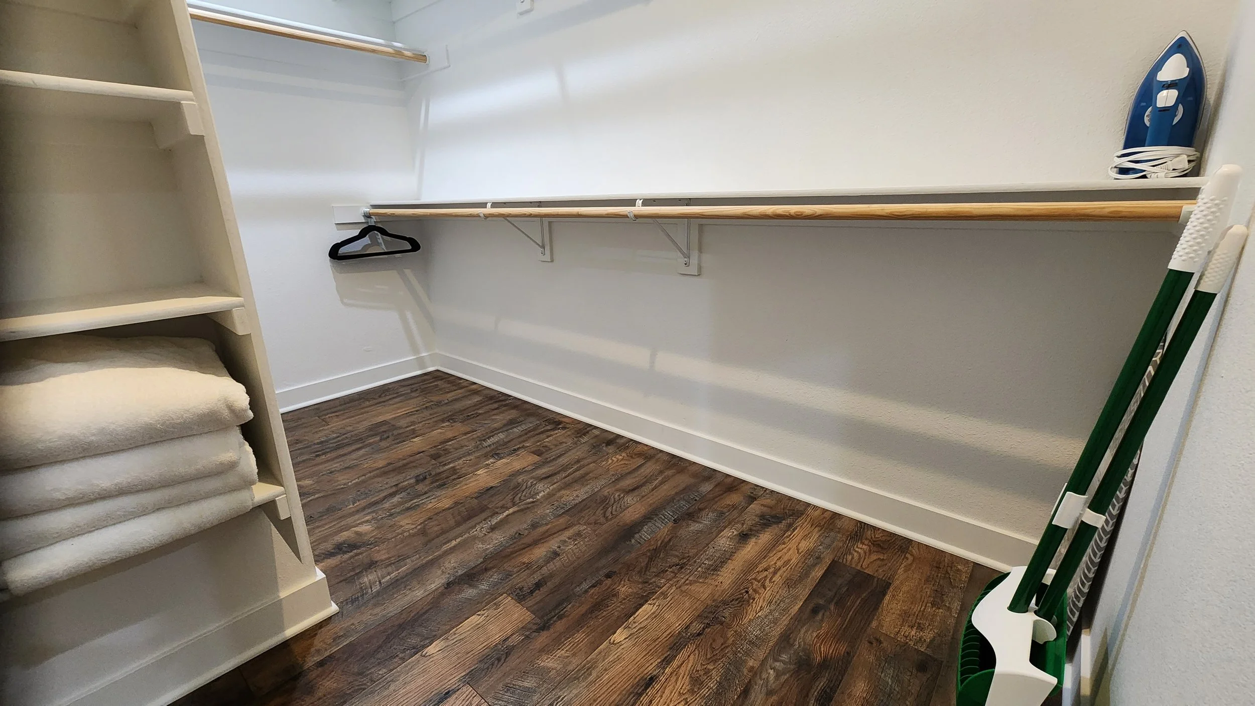 Empty walk-in closet with wooden flooring, white shelves with folded towels, built-in hanging rods, an iron, and cleaning tools including a broom and mop leaning against the wall.