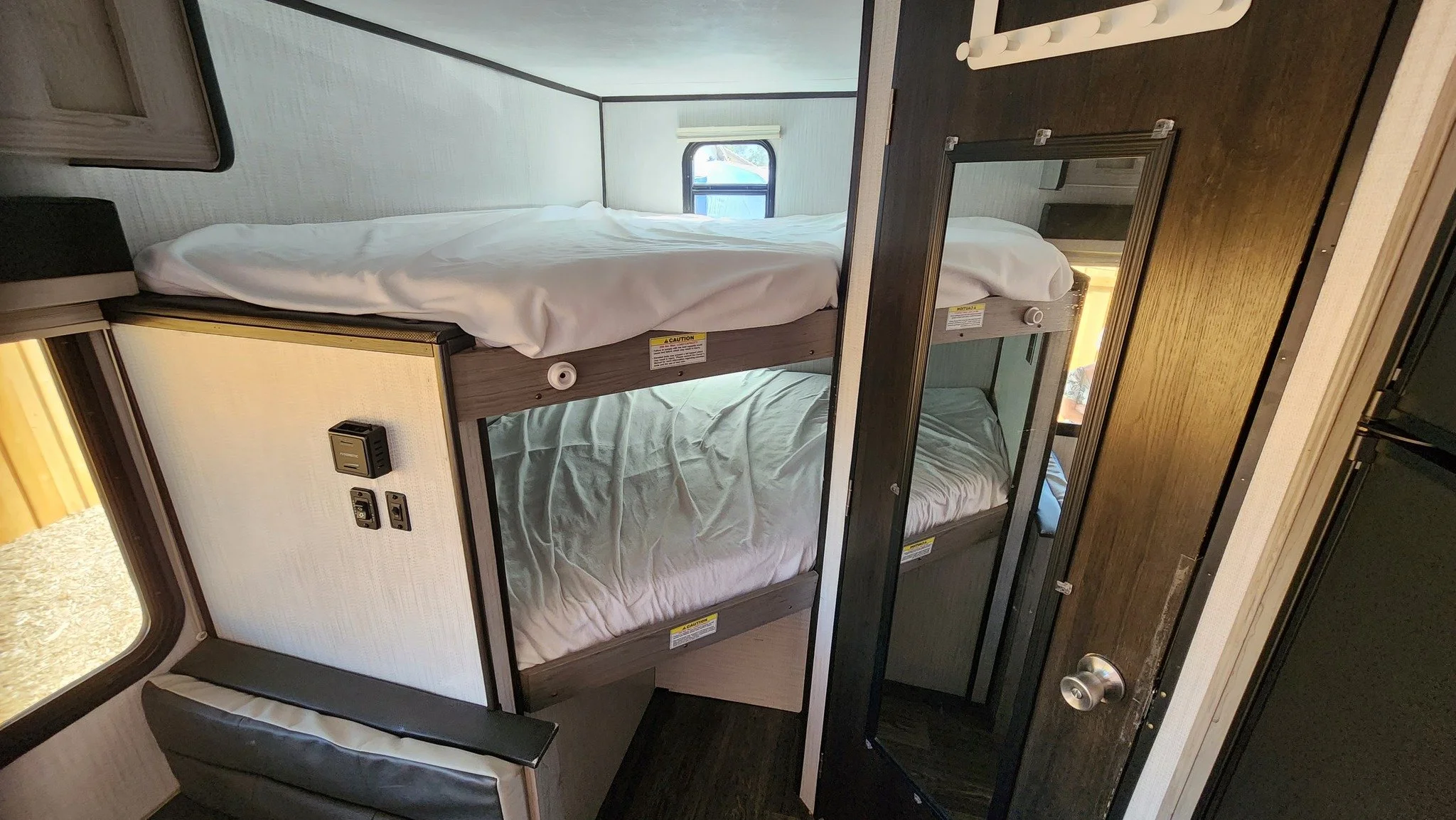 Interior of a compact camper with stacked bunk beds, small windows, a mirror, and a door.