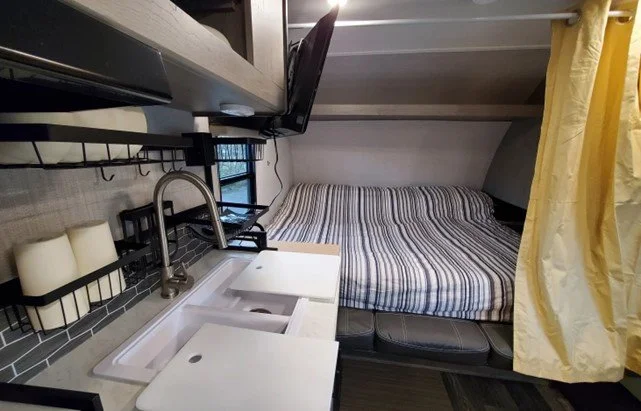 Compact kitchenette with white countertop and sink, black wire rack with toiletries, and a bed with striped bedding in a small, cozy room.