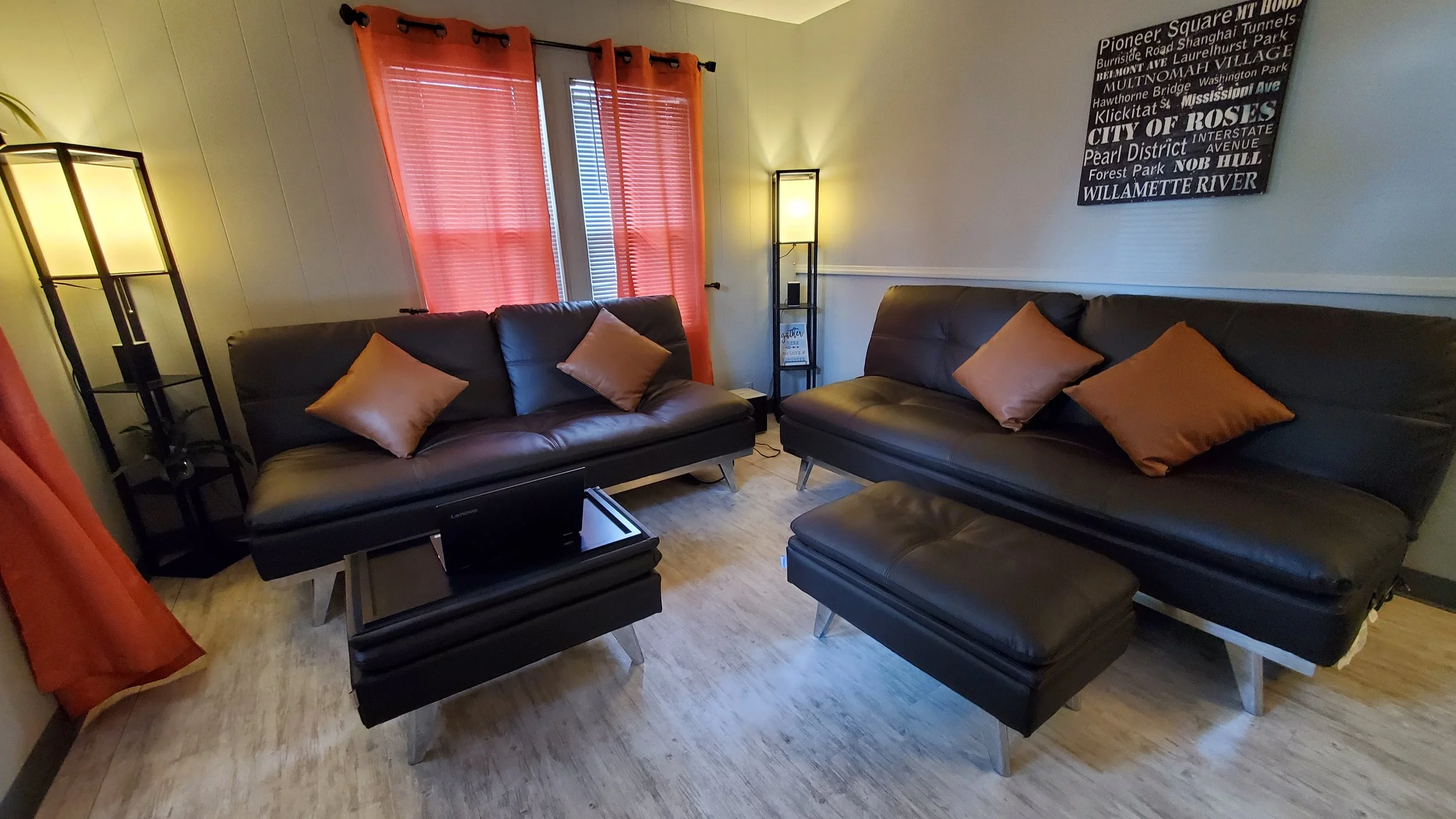Living room with two black leather sofas, orange throw pillows, orange curtains, a wooden coffee table with a closed laptop, a standing lamp, and framed art on the wall with neighborhood names.