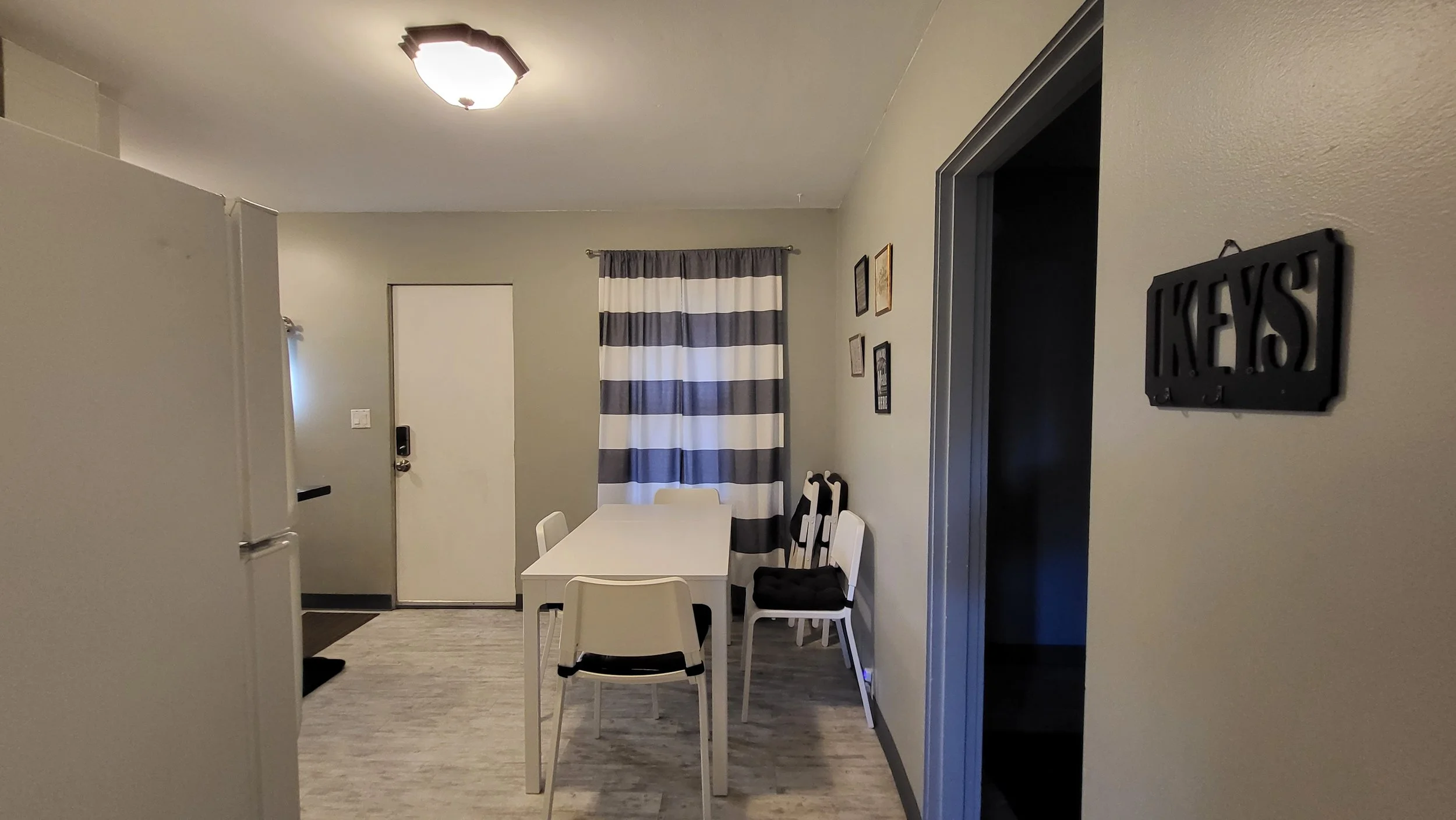 Interior view of a dining area with a white table and chairs, a striped curtain on a window, and portrait pictures on the wall. There is a front door with a keypad lock and a sign that reads 'KEYS' on the wall.
