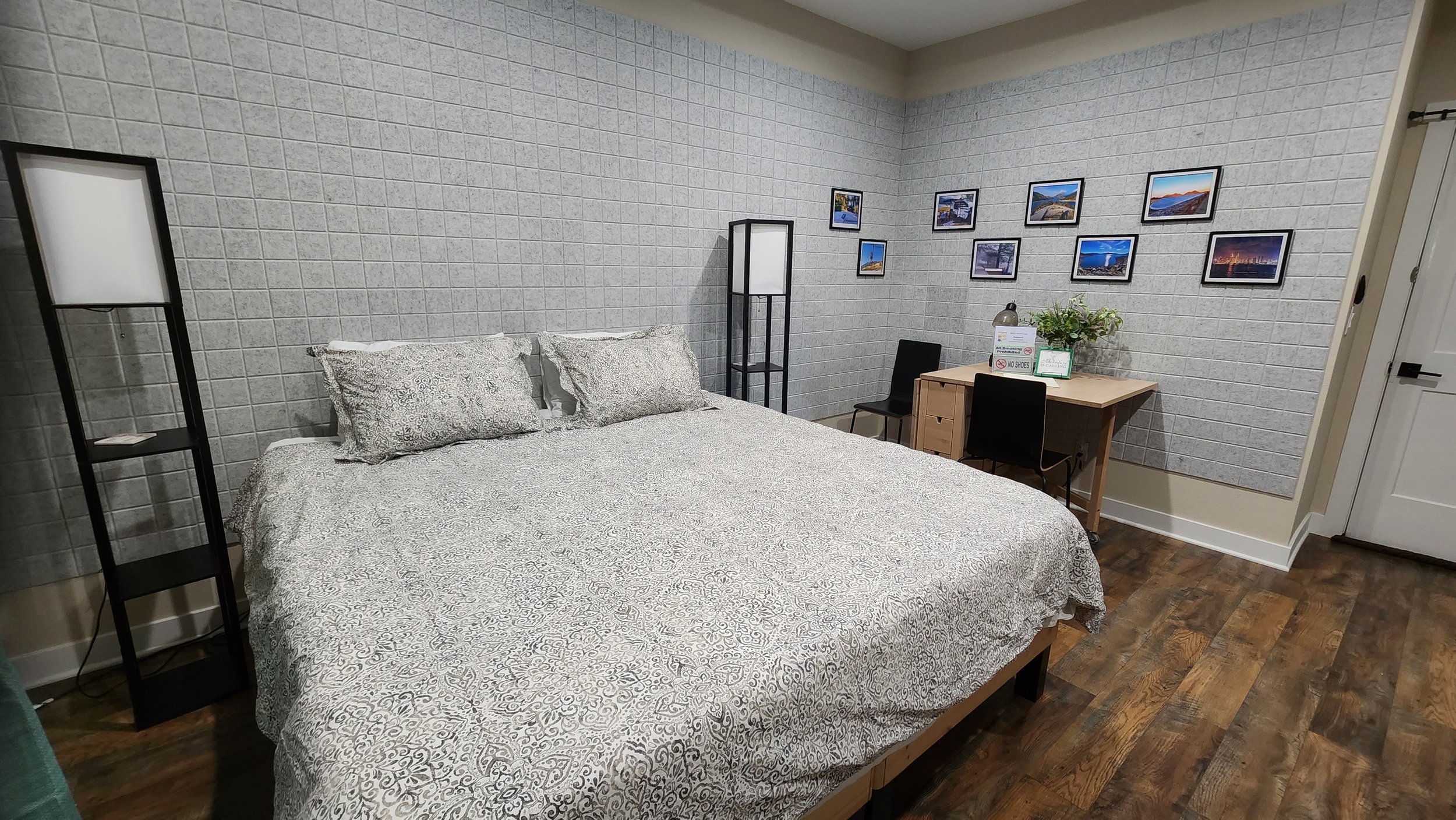 Interior of a bedroom with a bed, lamps, a desk with chairs, and framed landscape photos on the wall.