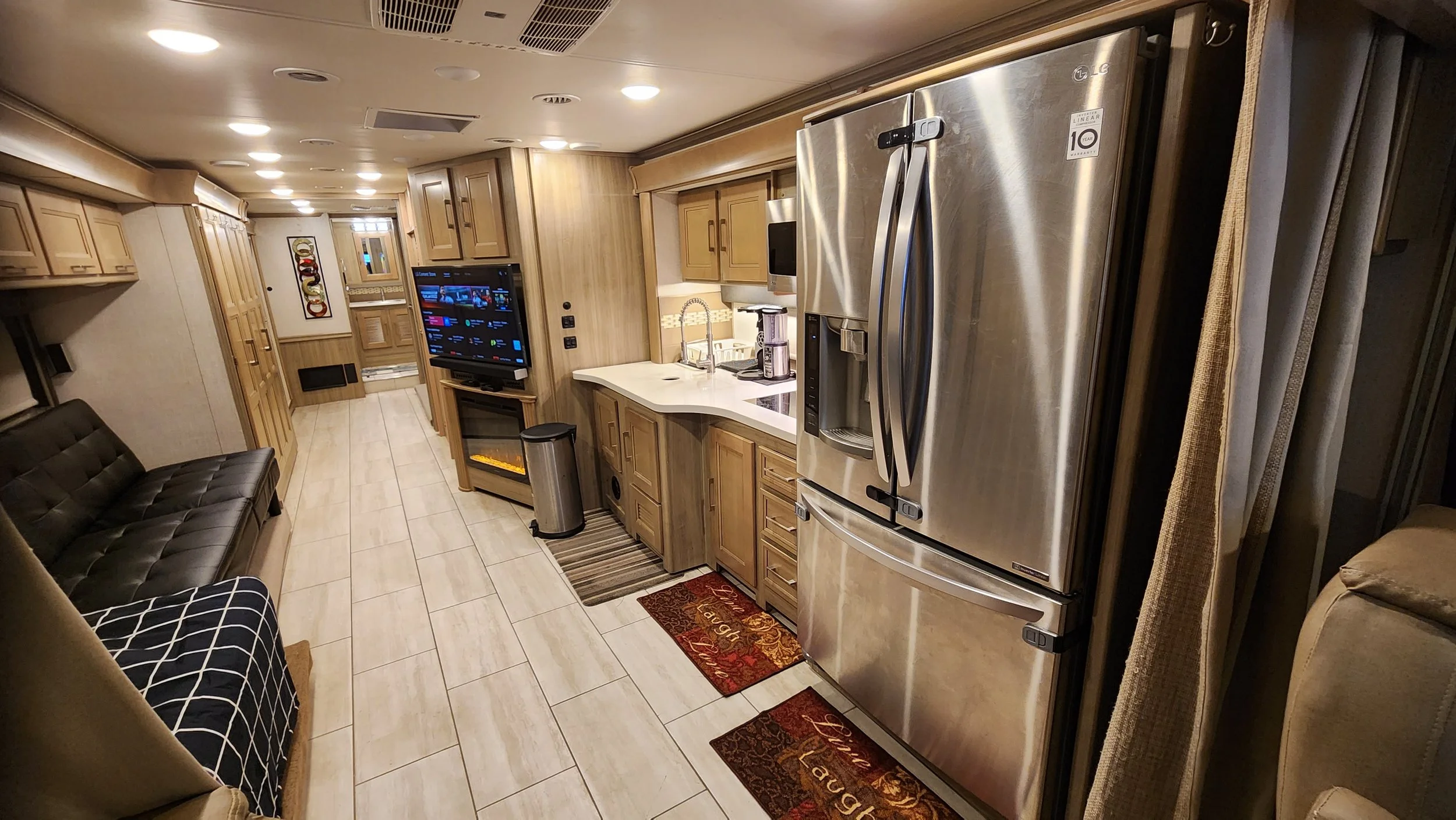 Interior view of a cozy RV or mobile home with a kitchen area featuring stainless steel appliances, wooden cabinets, a black leather sofa, a television, and various rugs on tiled flooring.