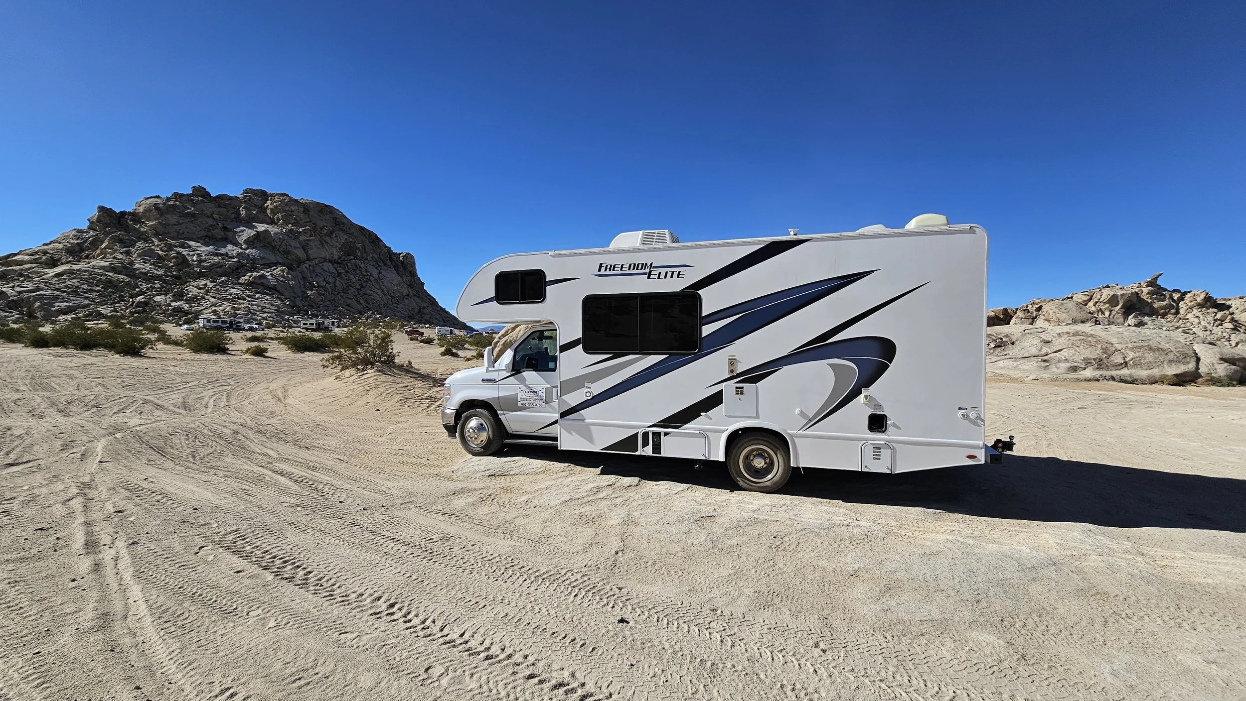 A white campervan with black and gray graphics parked on sandy desert terrain near rocky hills under a clear blue sky.