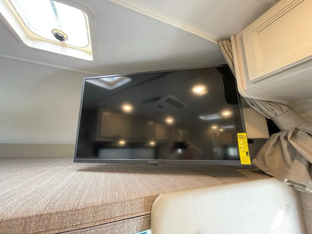 Flat-screen television on a beige bed or cushioned surface in a room with ceiling lights and a ceiling vent.