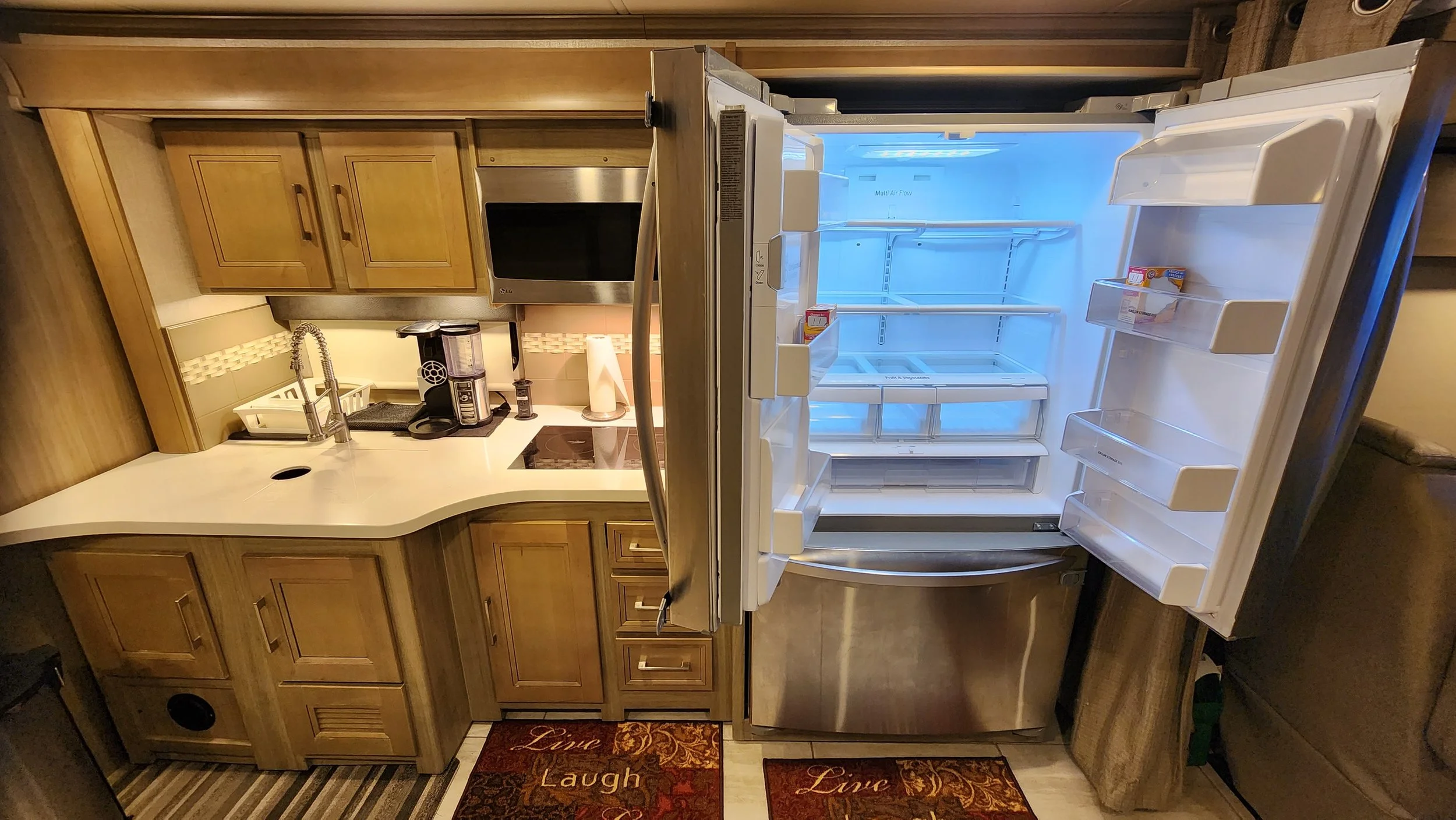 Open stainless steel refrigerator with empty shelves in a compact kitchen with wooden cabinets, a white countertop, a coffee maker, microwave, and sink.