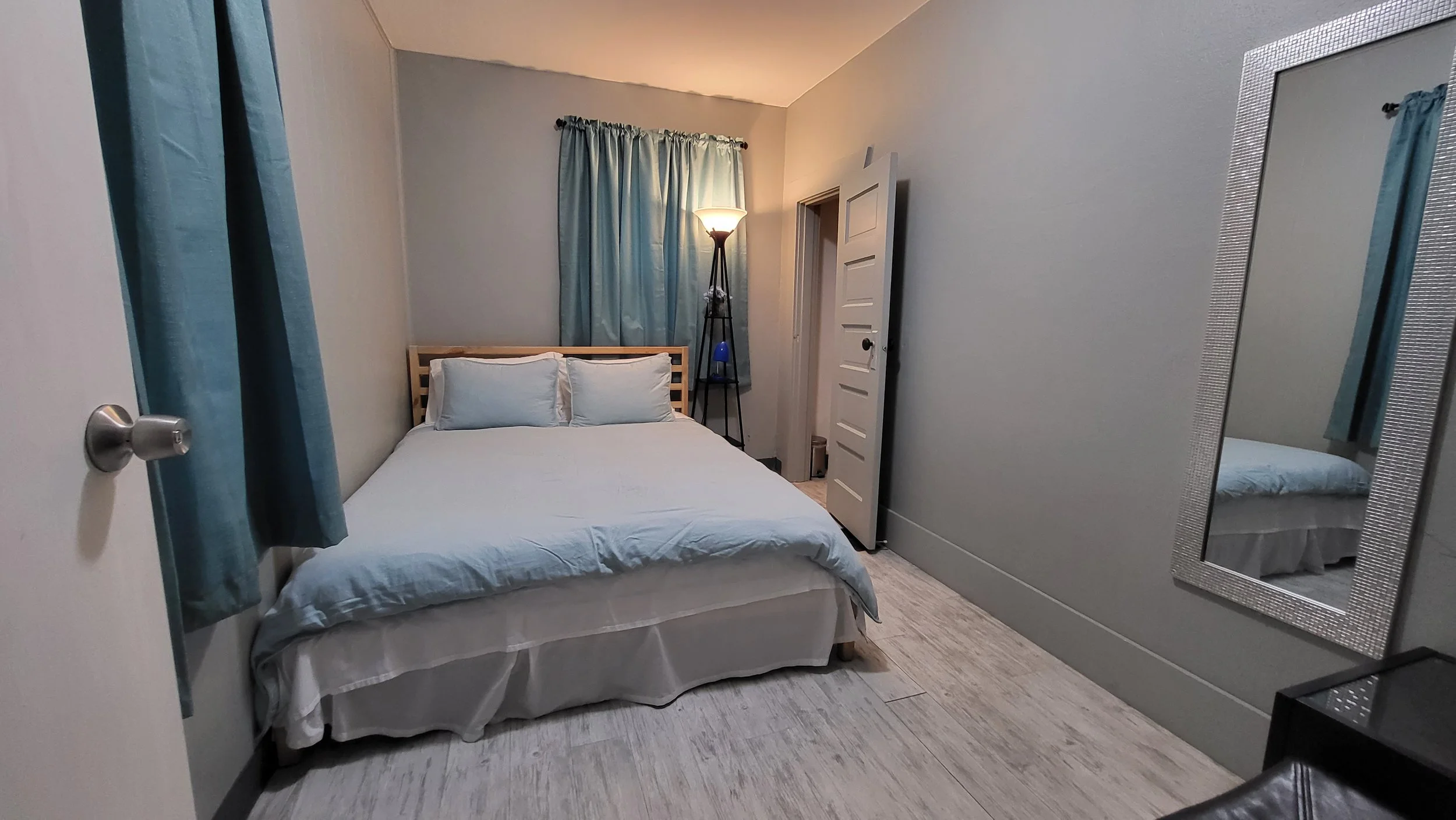 A small bedroom with a bed with white bedding and pillows, blue curtains, a standing lamp, a mirror on the wall, and a door slightly open.