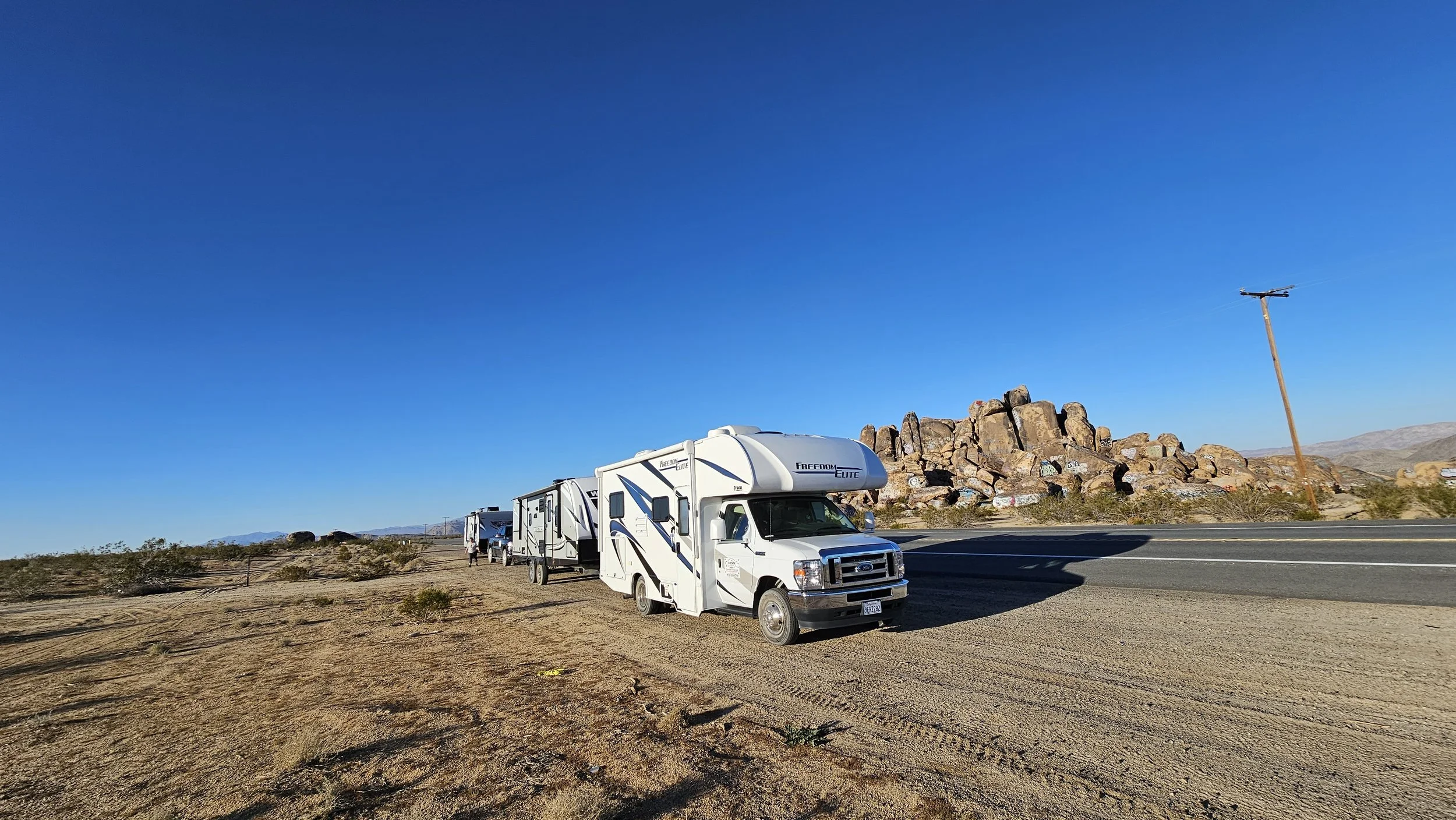 Multiple RVs parked on a dirt area next to a road in a desert landscape with large rocks and a clear blue sky.