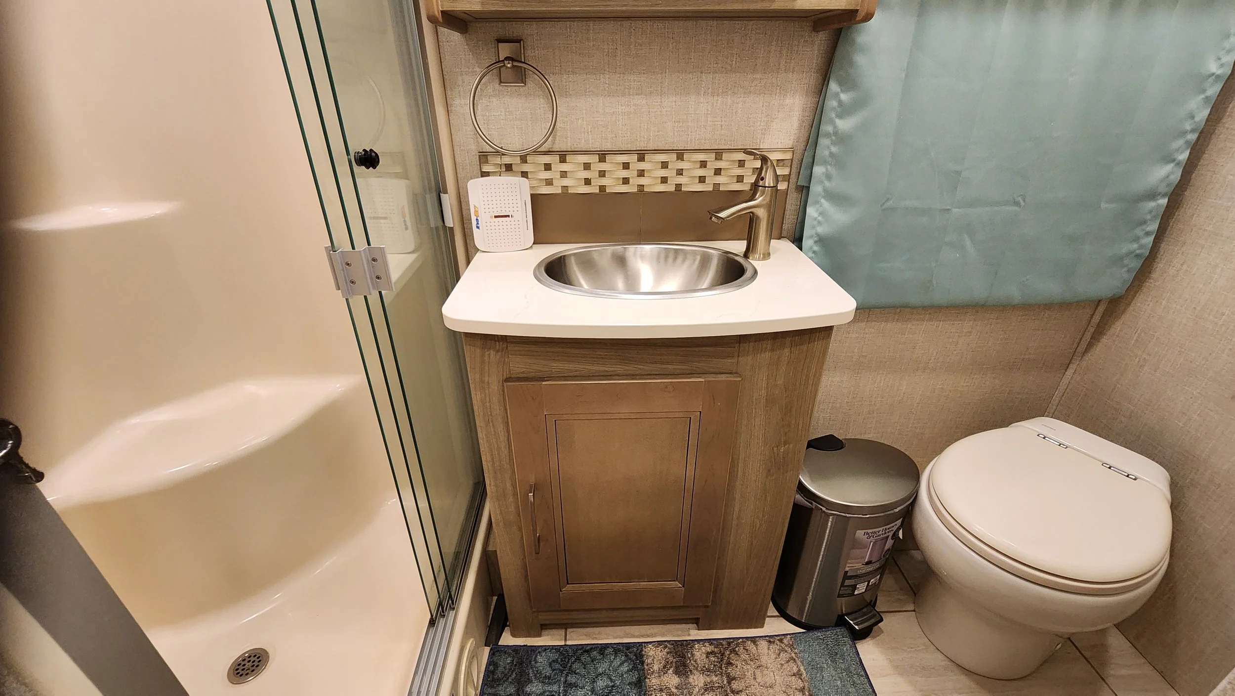 Bathroom with shower, small wooden cabinet with a sink, a trash can, a toilet, mirror, and light blue privacy curtain.