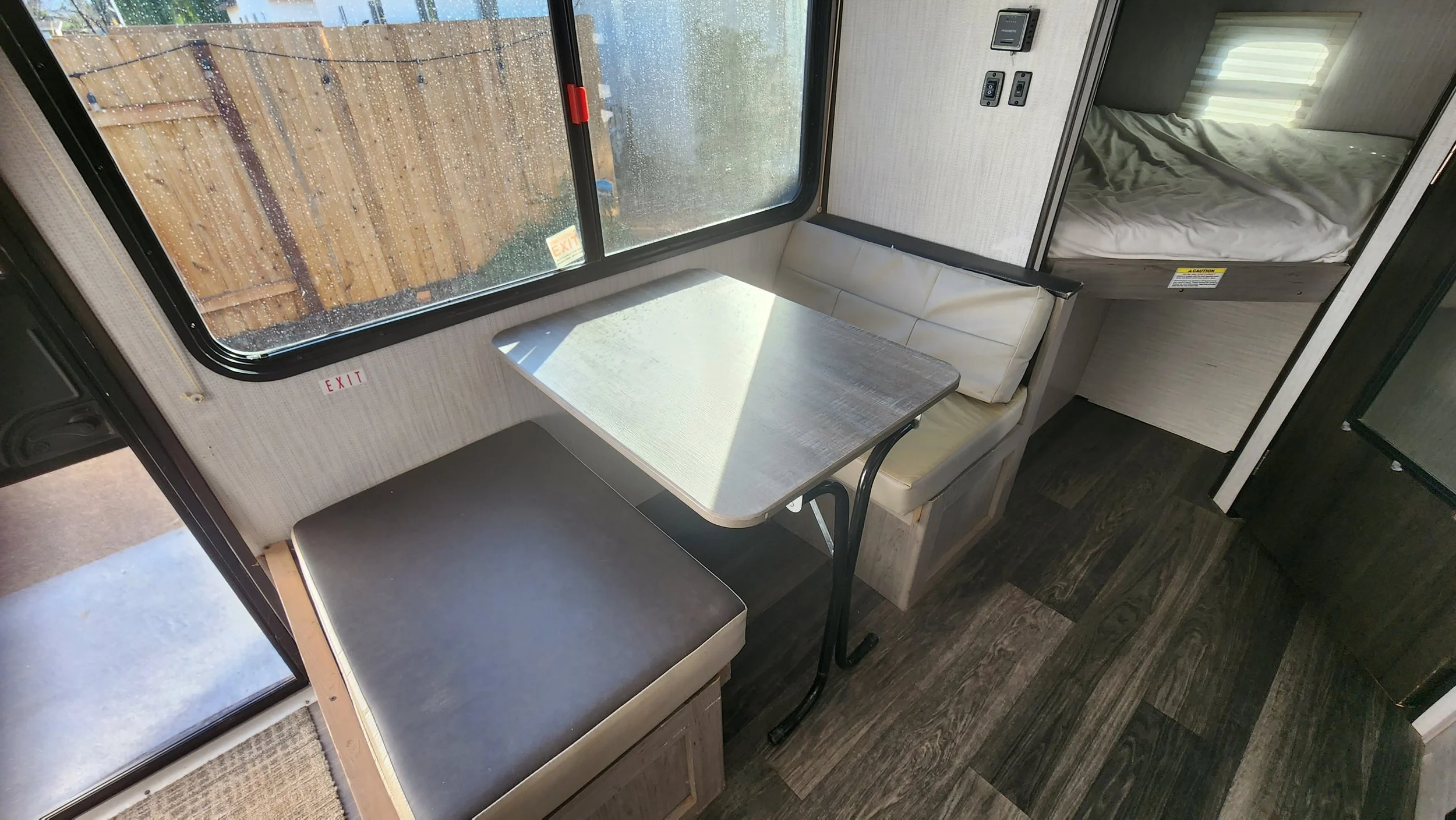 Inside a RV, a small dining area with a table, cushioned bench seats, a window with rain droplets, and a sleeping area with a bed.
