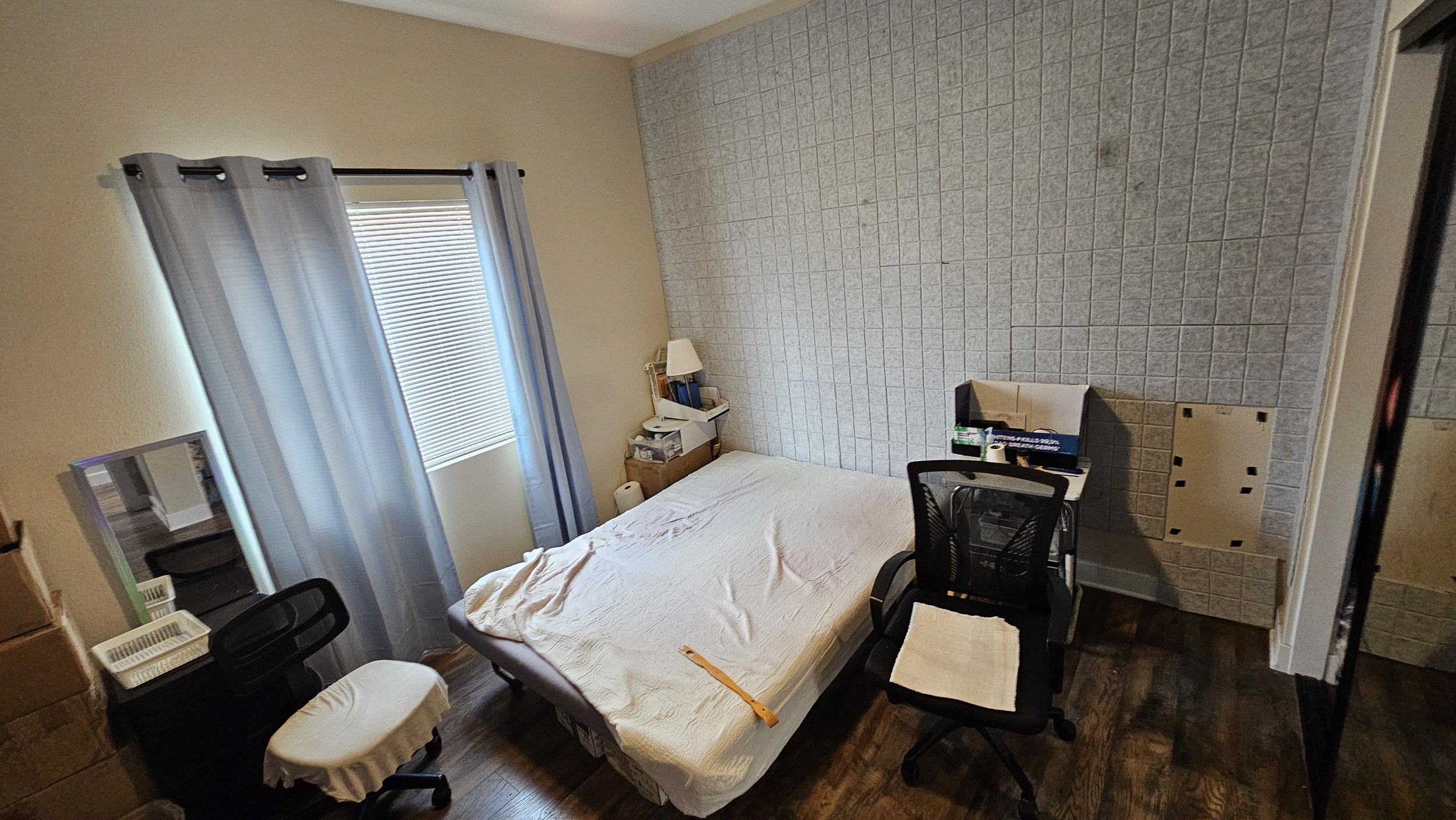 A bedroom with a bed, a window with blinds, and various furniture and boxes, with a wood floor and a tiled wall.