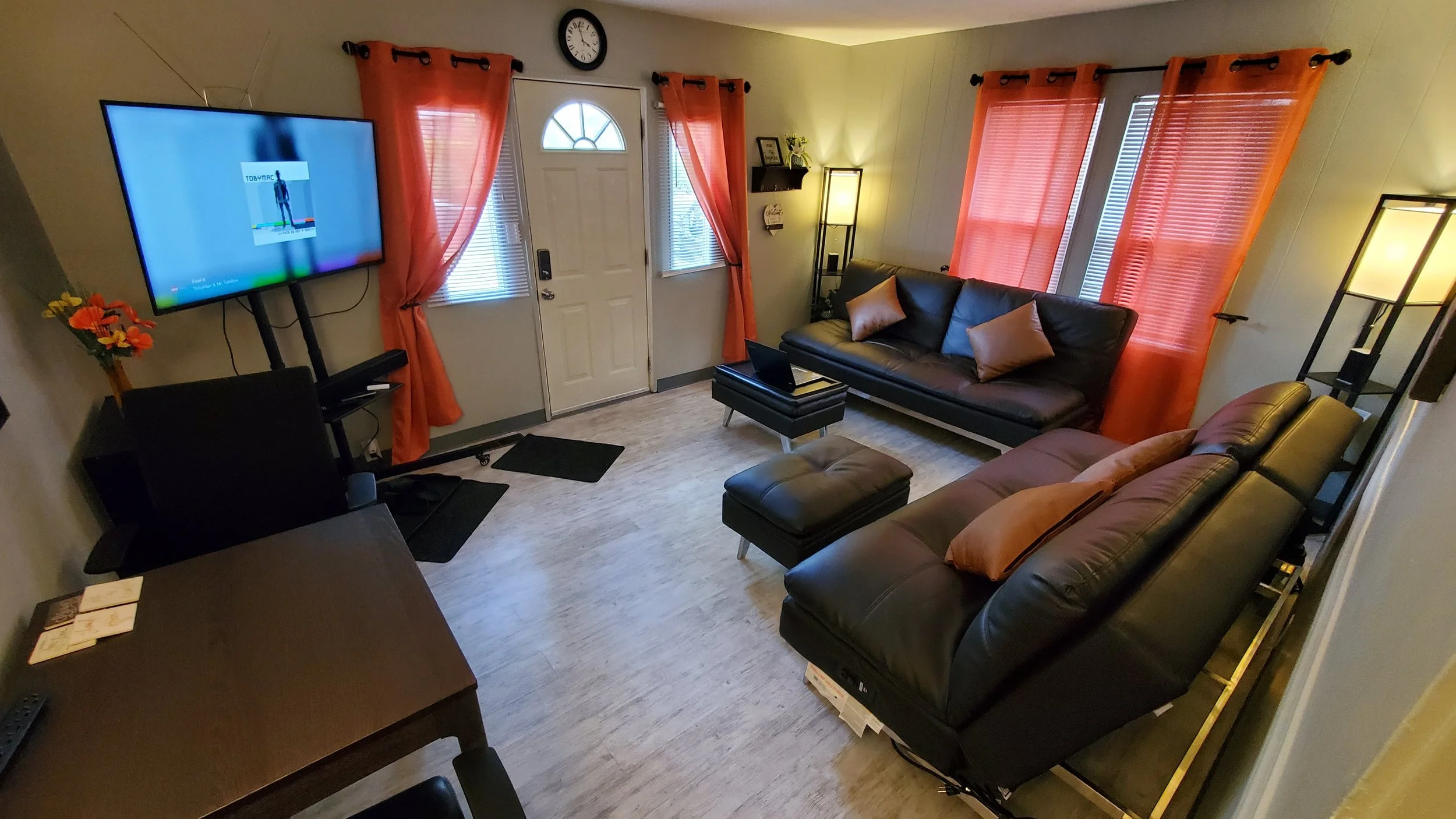 A living room with beige walls, a white front door, and five windows covered with red curtains. There is a large black leather sofa with pillows, a matching ottoman, a small black side table with a laptop, a coat rack with a lamp and a shelf, a wall clock, and a flat-screen TV on a stand. The floor is light-colored wood or laminate.