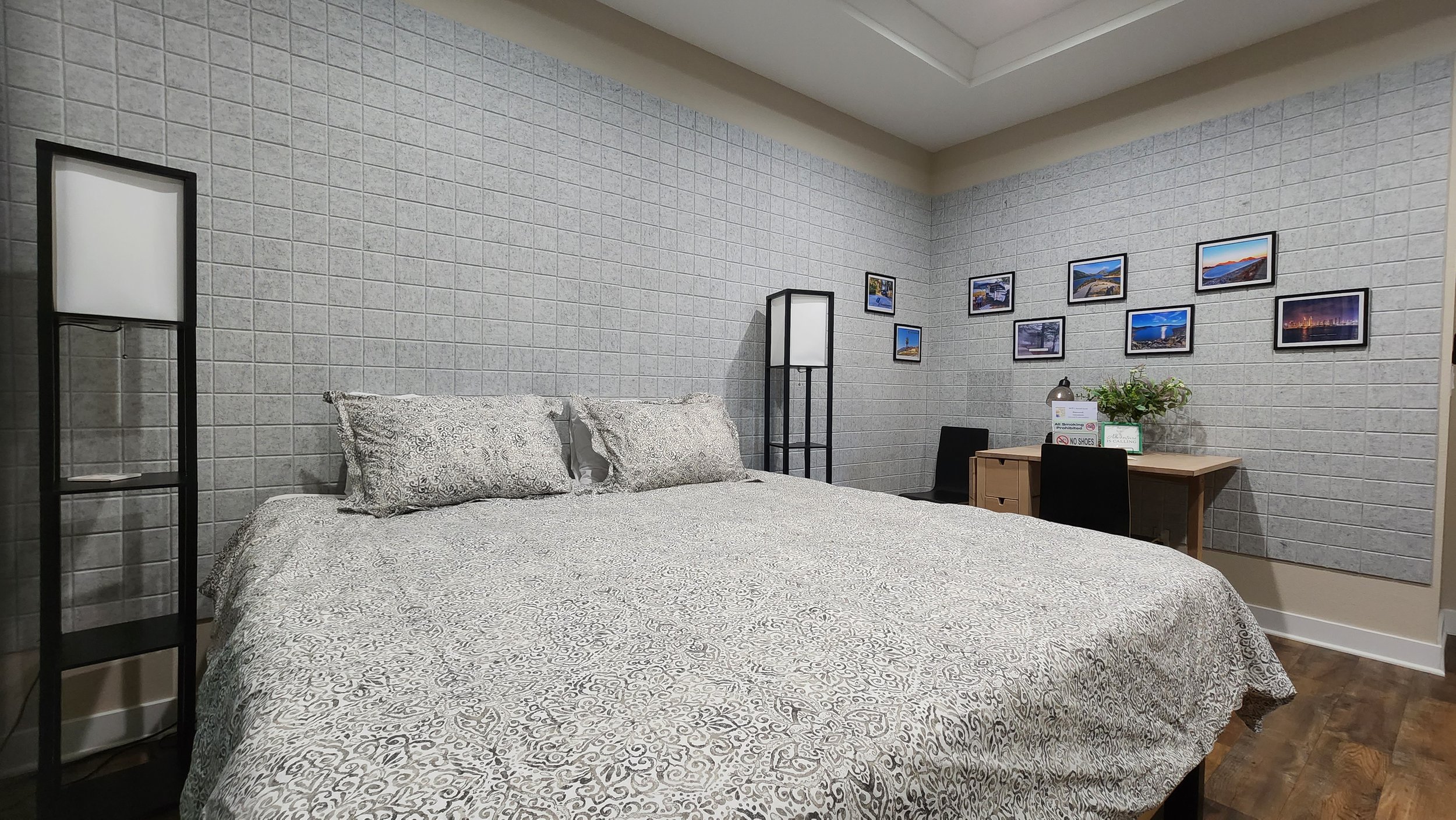 Hotel room with a large bed, two tall floor lamps, a small desk with a plant, framed photographs on the wall, and wooden flooring.