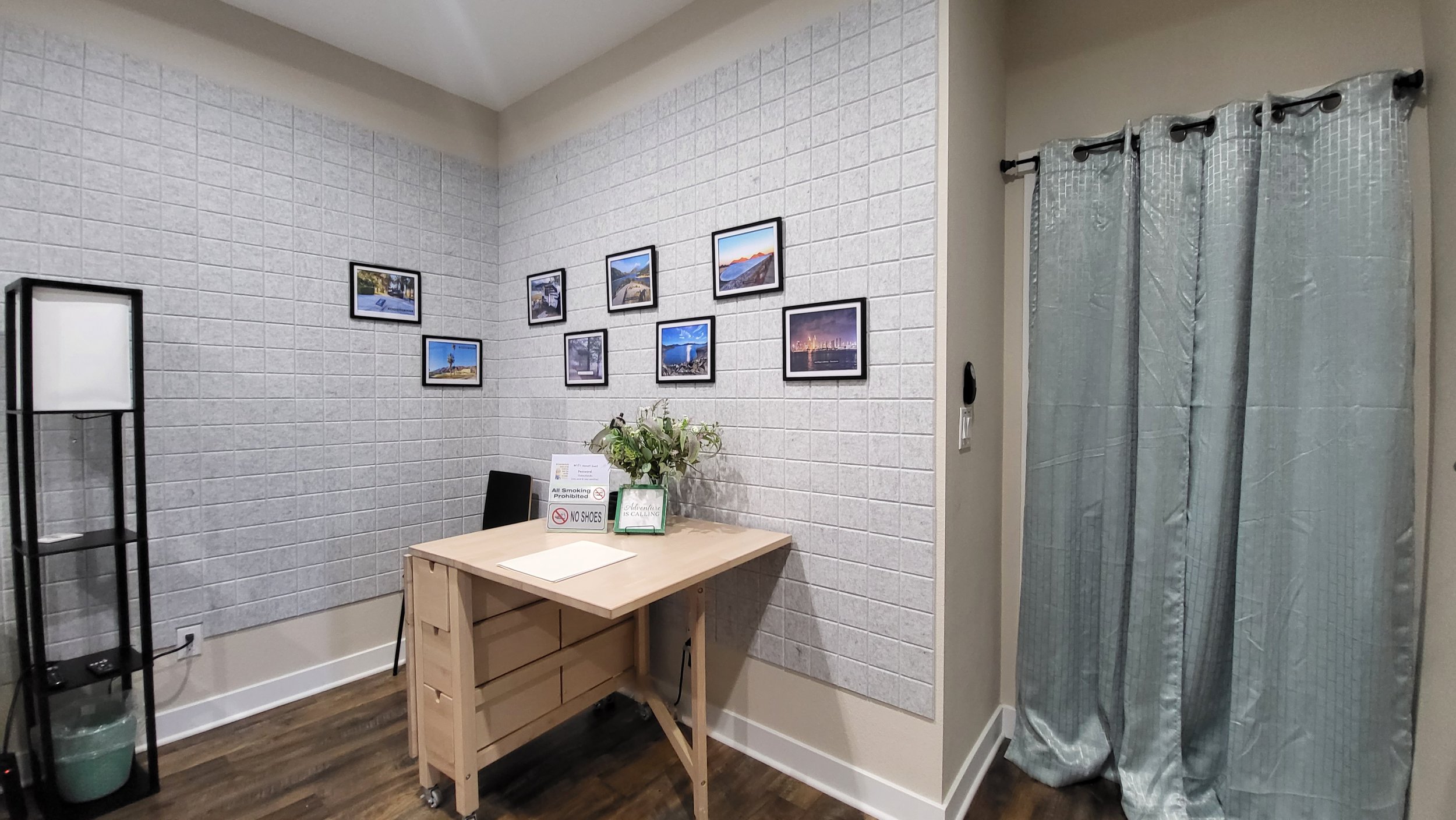 A small room with a light wood table, a black chair, a bouquet of flowers in a green vase, and a photo collage on the wall. There is a sign on the table and a tall black shelf with a lamp and a green bucket. A curtain covers an opening on the right s