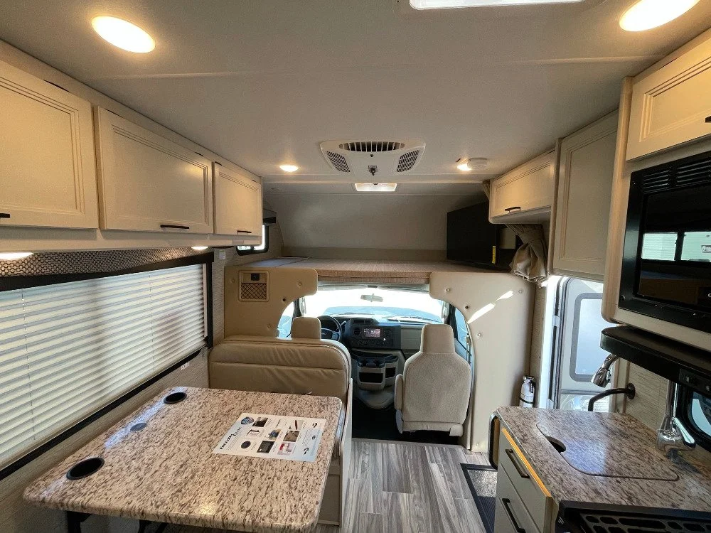 Interior of a motorhome or RV with a small dining area, kitchenette, and driver's cabin visible at the front.