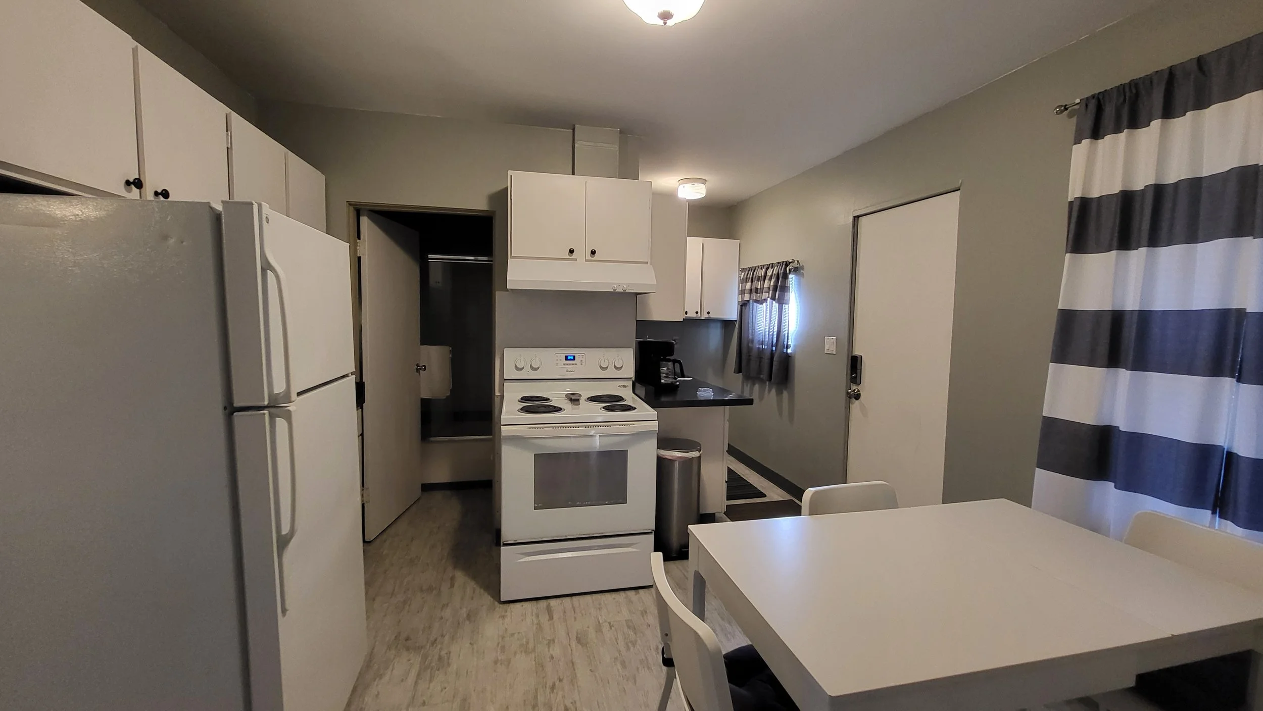 Kitchen with white refrigerator, stove, black countertop, coffee maker, window with plaid curtains, and a white table with chairs.