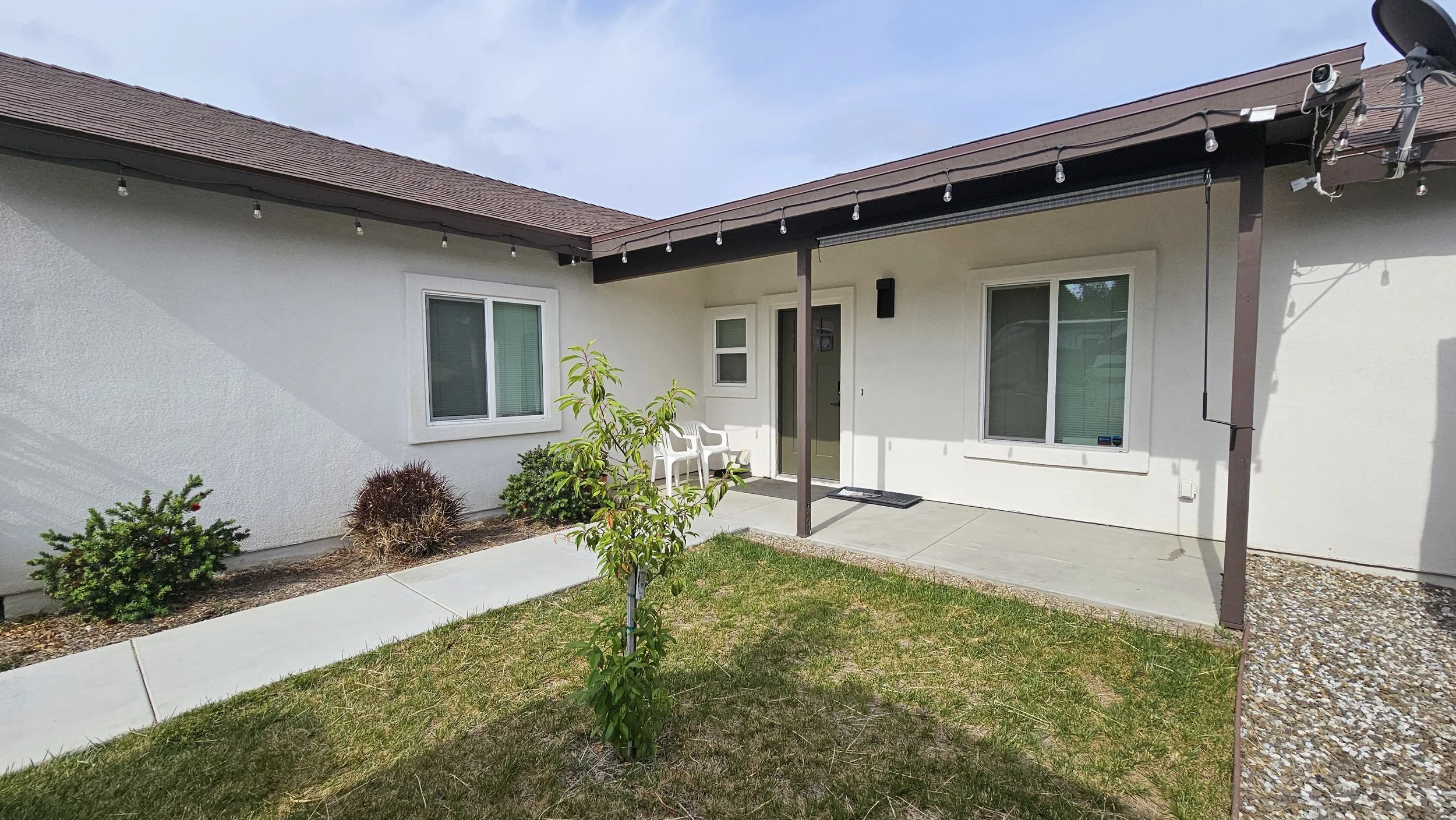 Backyard patio with a small grass lawn, a concrete walkway, two white plastic chairs near the door, and a white house with two windows, string lights hanging from the roof, and a small green tree in the yard.