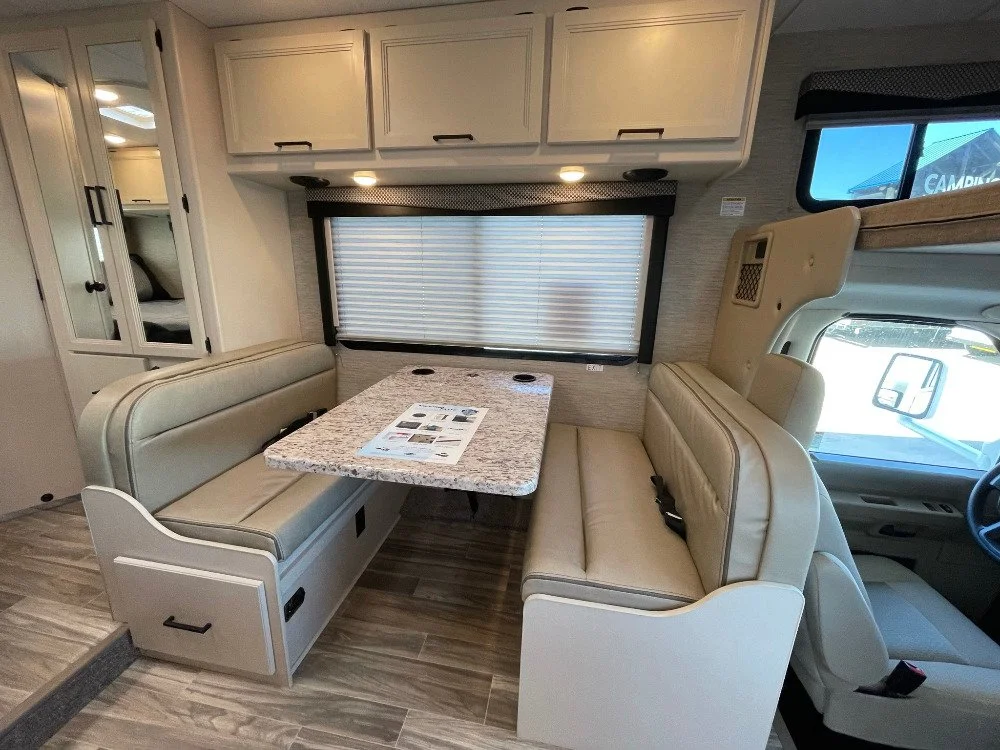 Interior view of an RV with a dinette area, featuring a granite-like table, beige cushioned bench seats, overhead cabinets, and window with blinds.