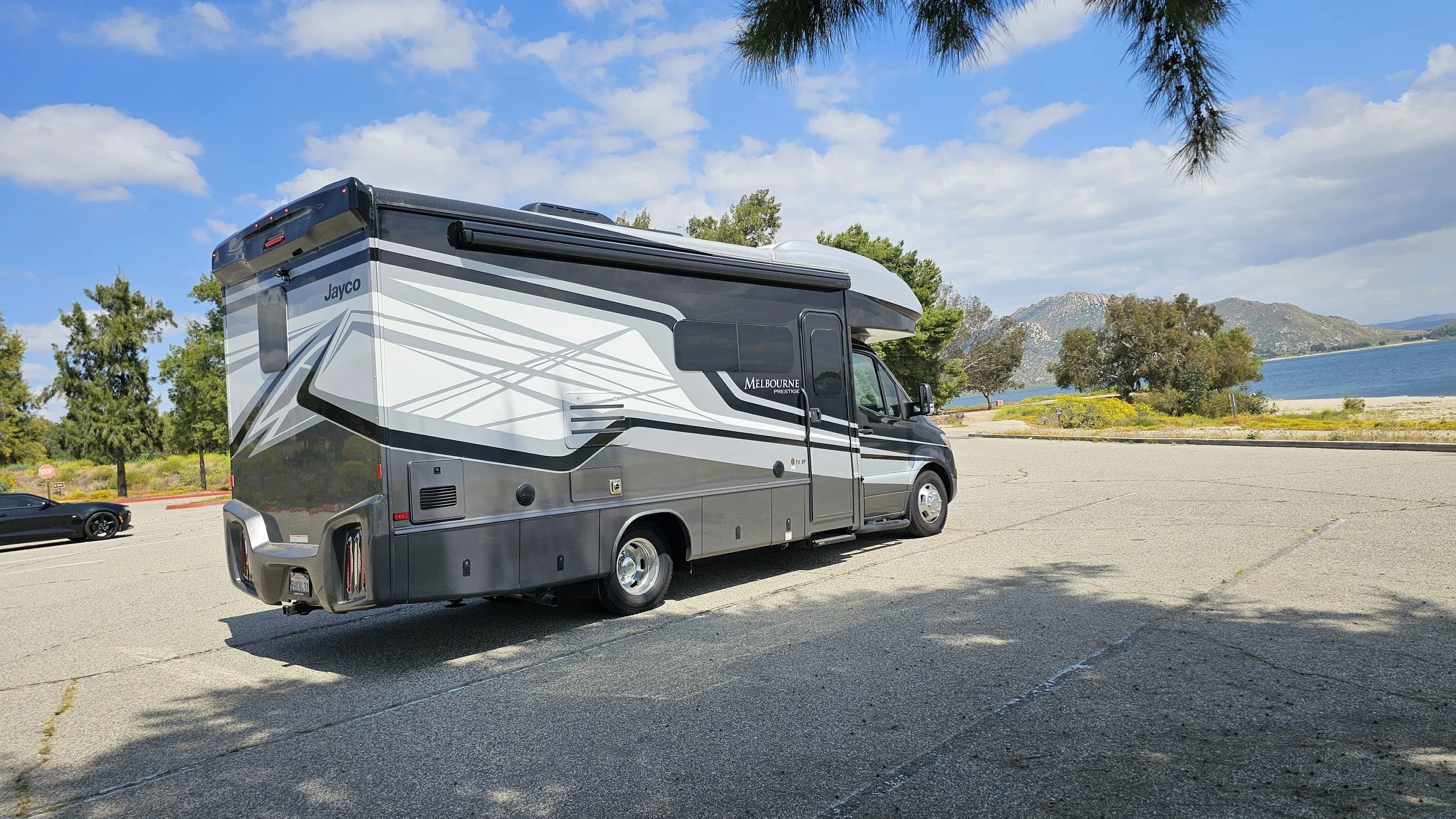 A black and gray motorhome parked near a lake with mountains in the background, trees, and a blue sky with clouds.