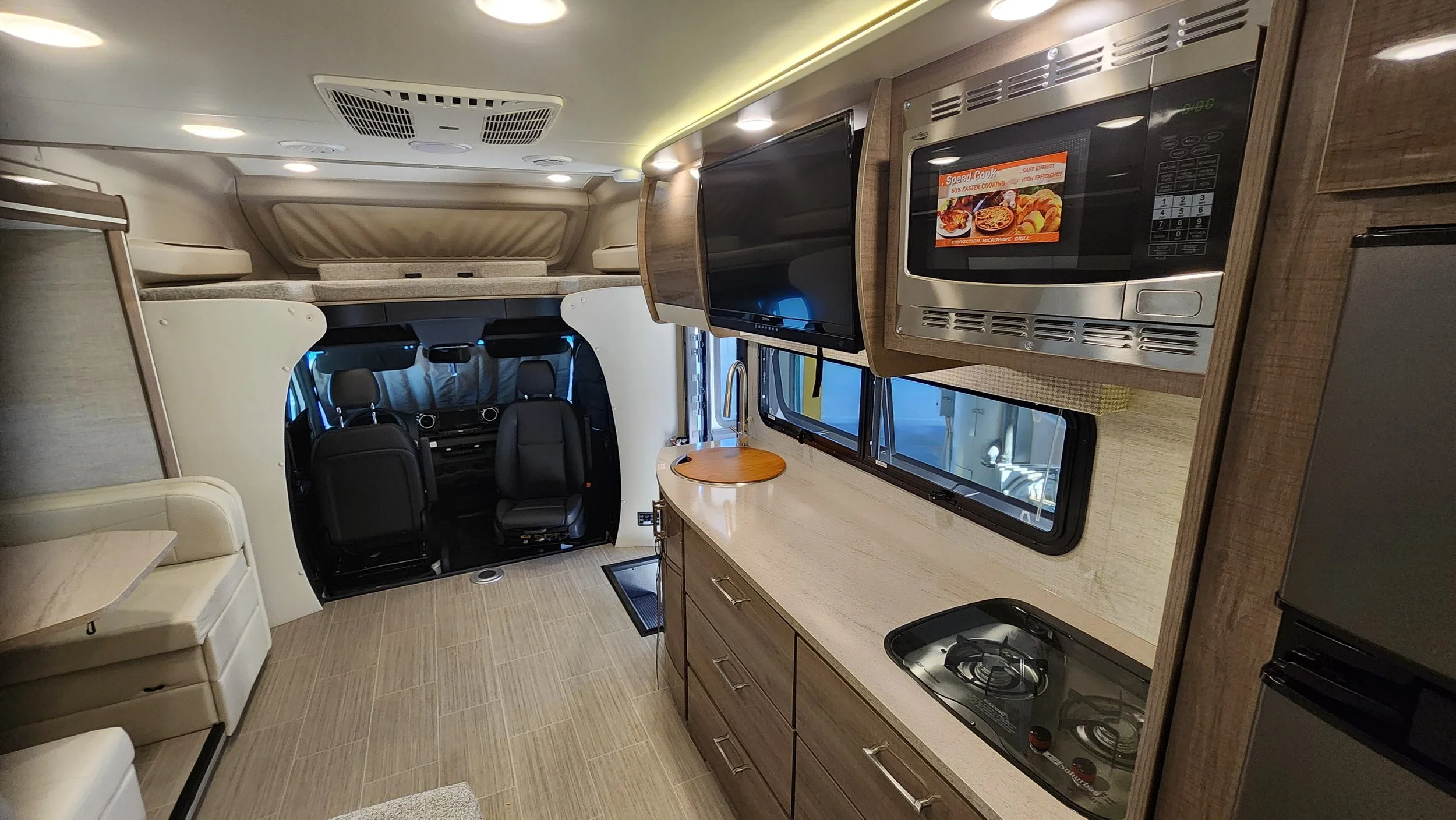 Interior of a camper van with a compact kitchen, microwave, TV, seating area, and driver cabin.