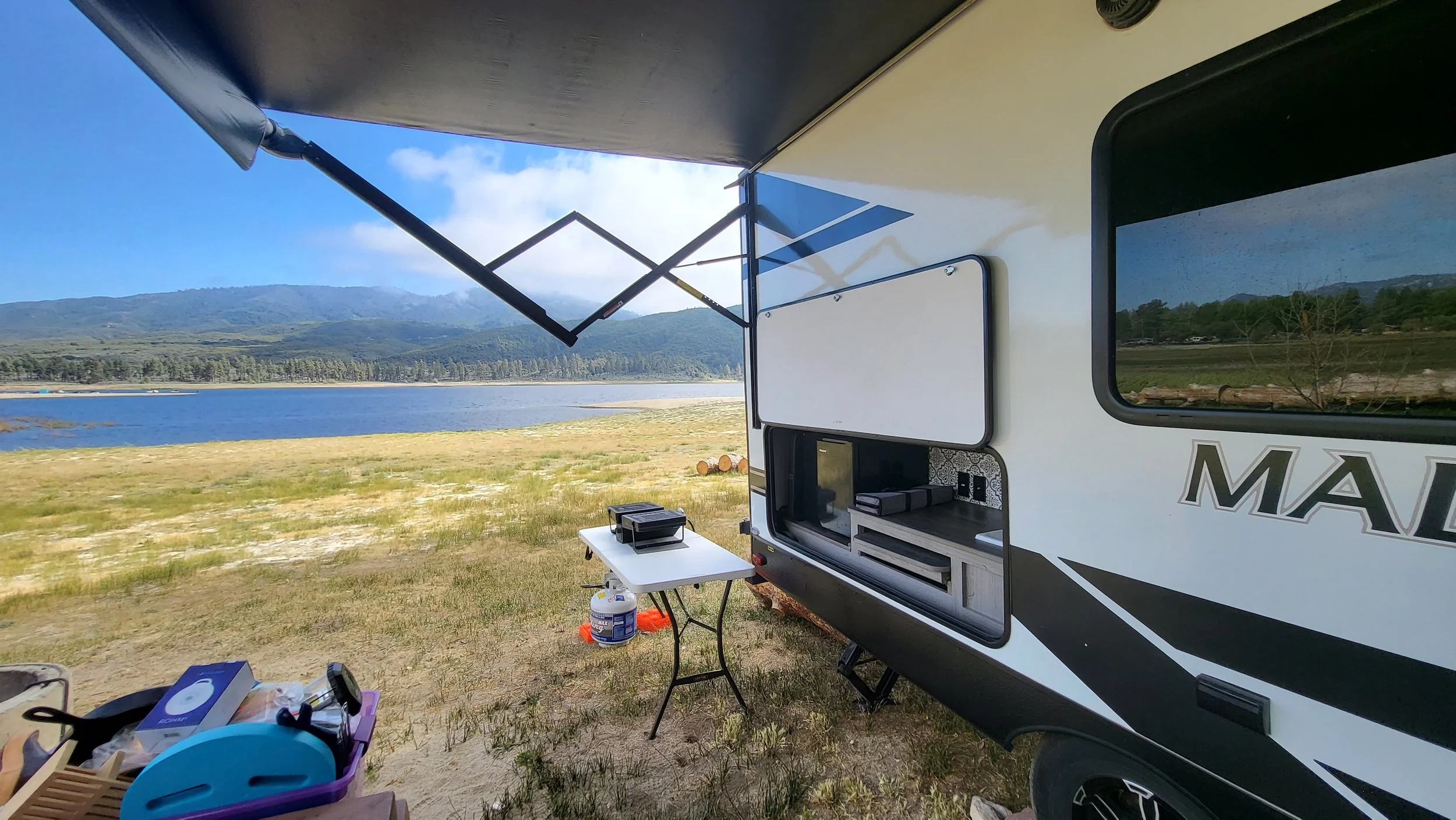 View of a camper trailer parked near a lake with mountains in the background. The slide-out of the trailer is extended, and a small table with some items is set up outside. The scene is sunny and peaceful.