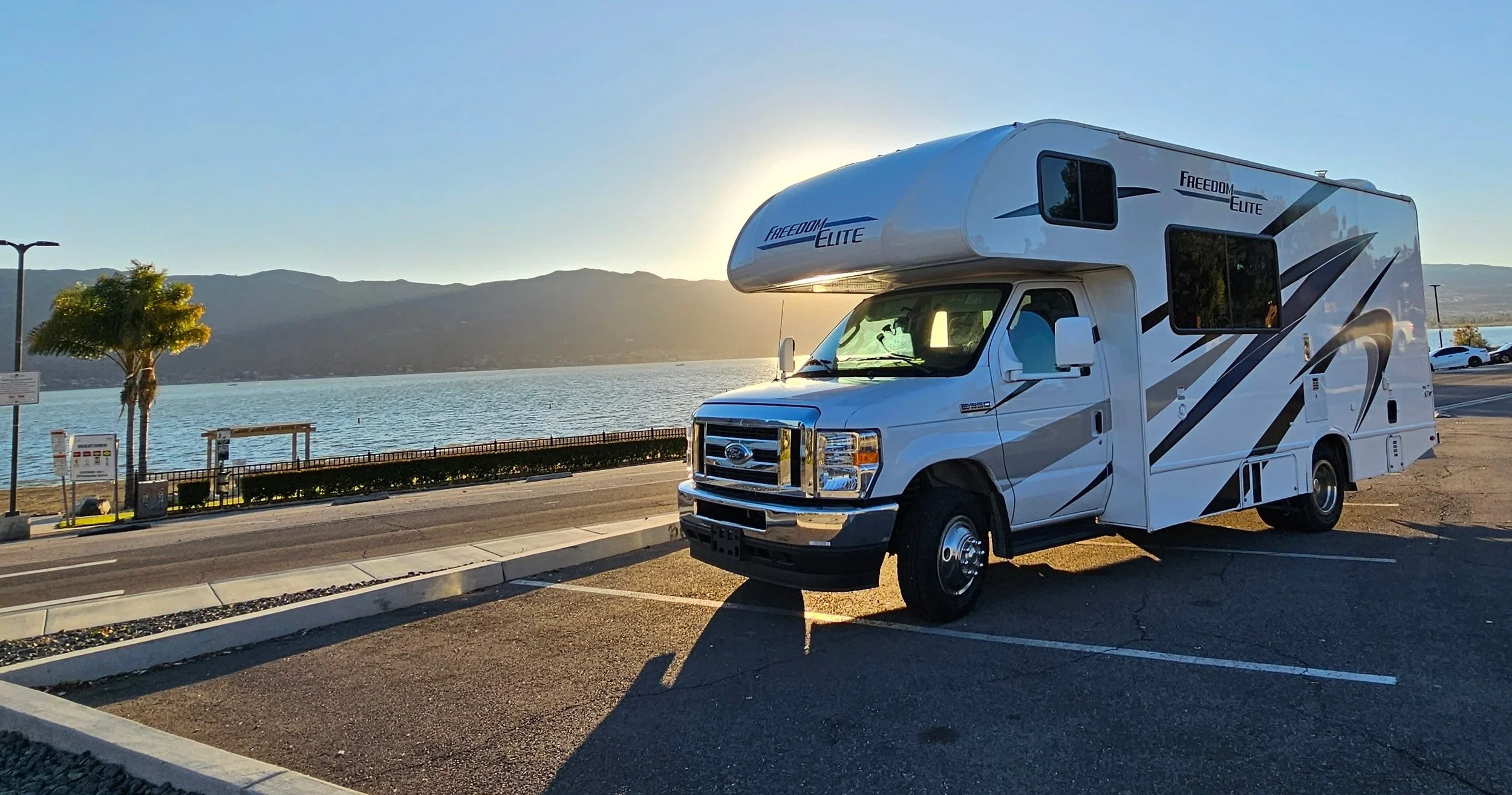 A white recreational vehicle (RV) parked near a lake with mountains in the background during sunset.
