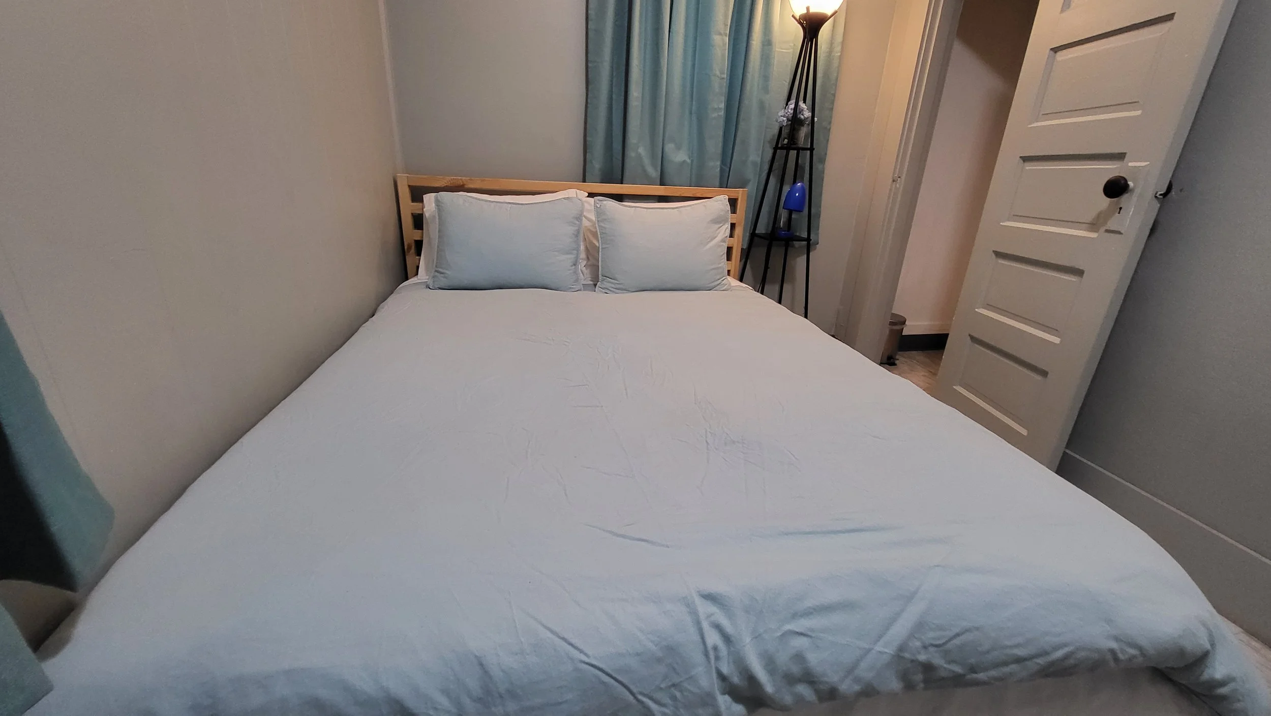 A neatly made bed with white bedding and three pillows in a small bedroom, with a standing black lamp next to a blue curtain and white louvered door.